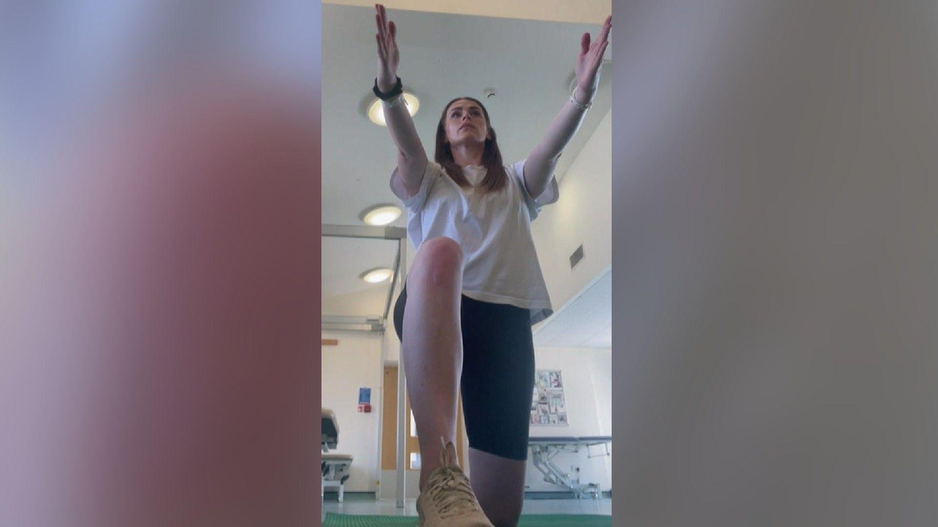 Emma Watt in a hospital lifting up one knee with her arms outstretched in front of her.