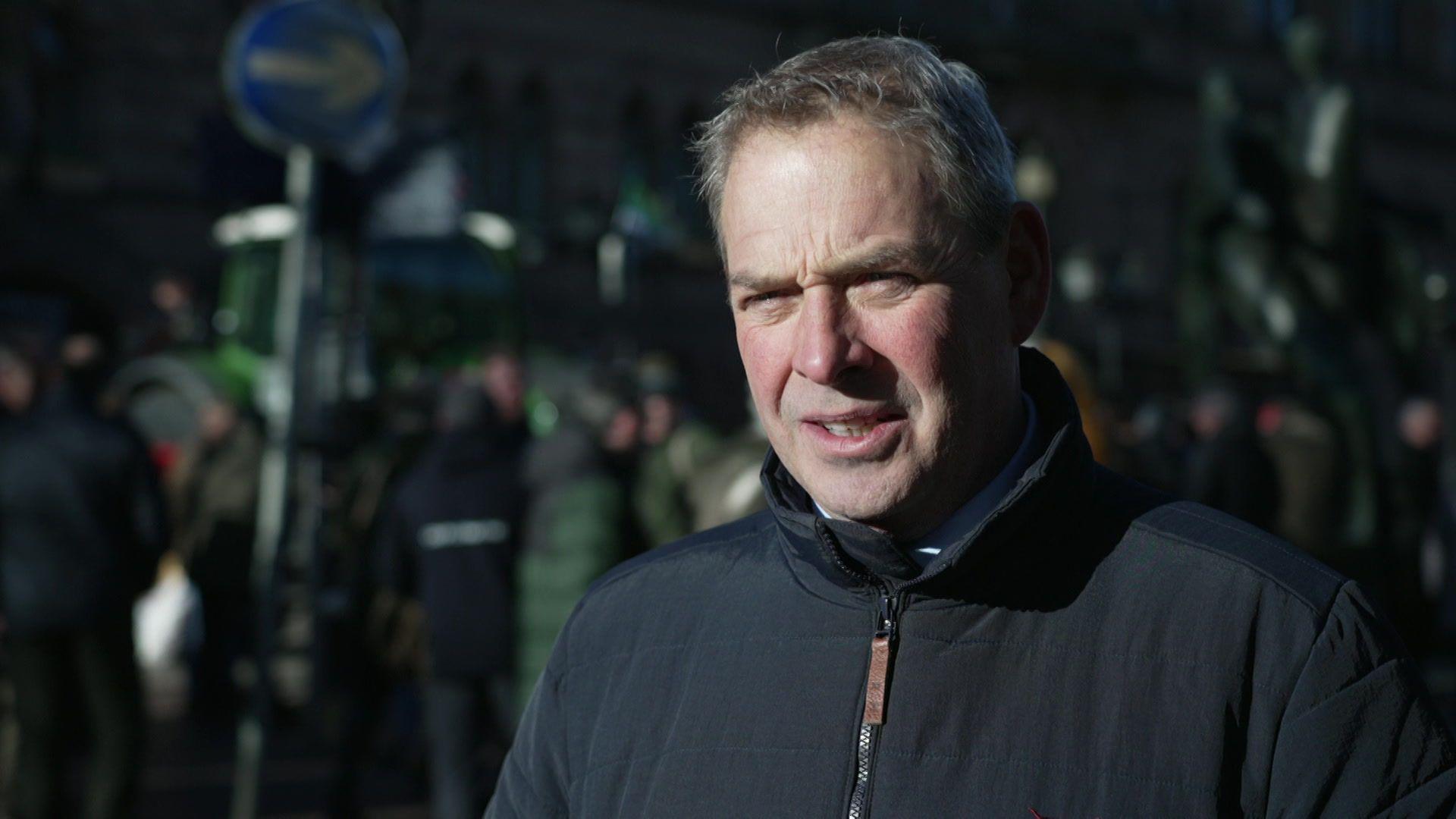 Richard Blackburn has short greying hair and is wearing a dark blue or black coat. A number of people and a tractor can be seen blurred out in Town Hall Square, Chester, in the background.