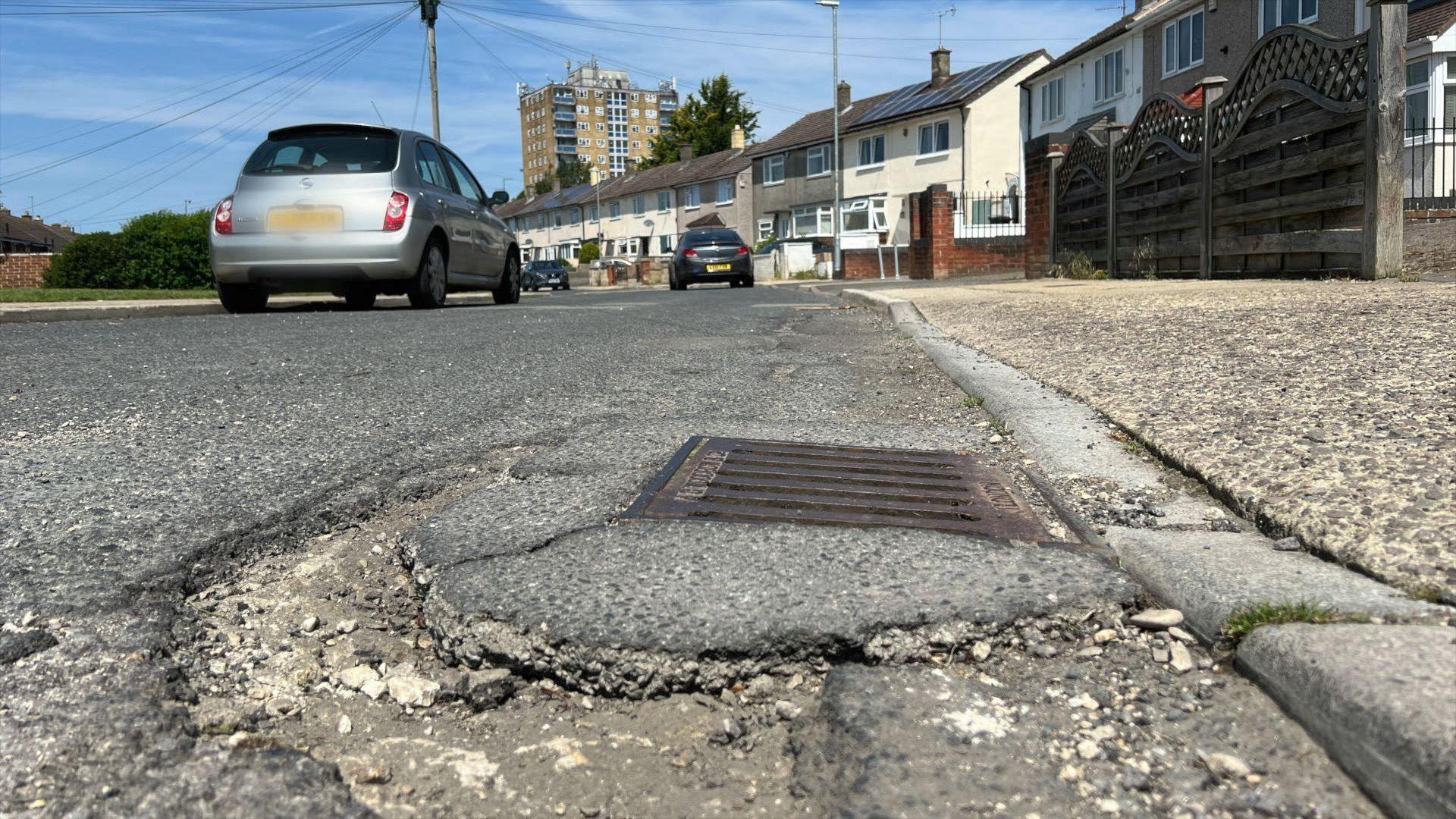 A pothole and crumbling pavement on a street of 1960s council houses, with one of Penhill's tower blocks in the background.
