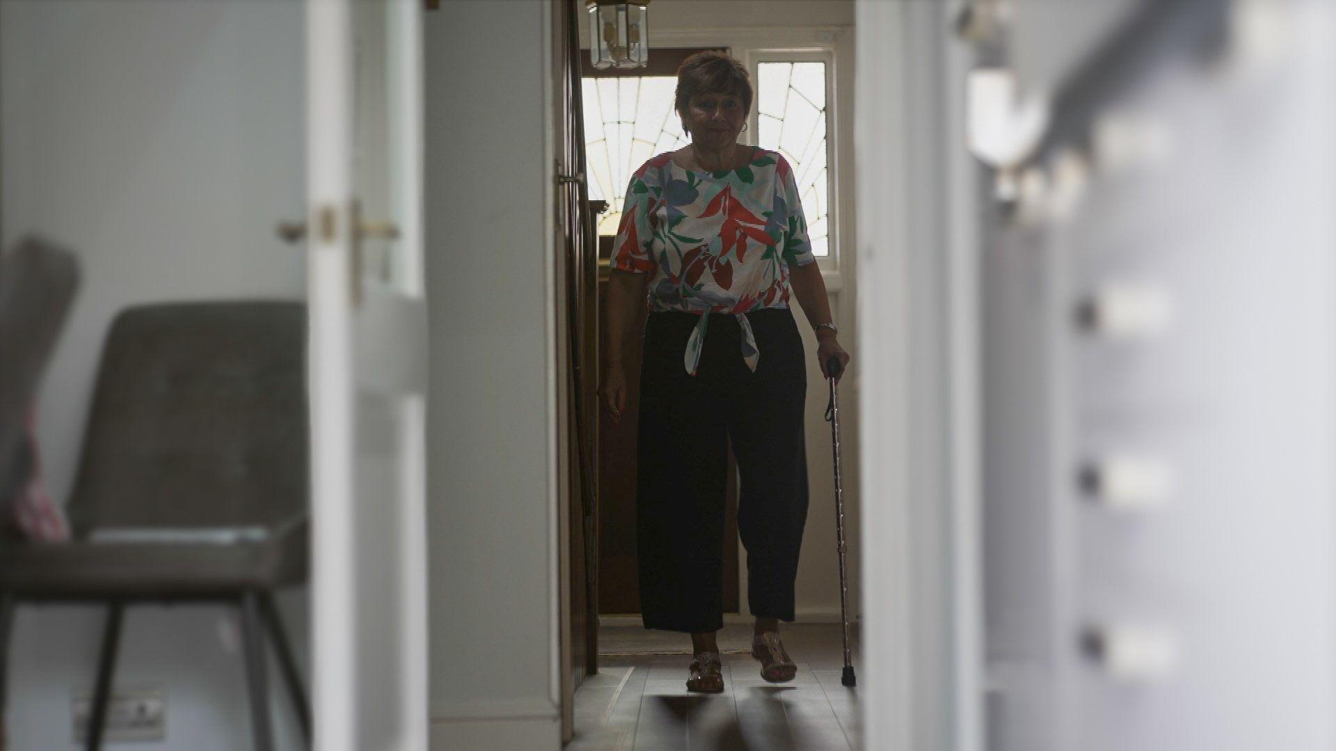 Guiliana walking inside her home, she is using a walking stick for support and has a floral top on and black trousers.