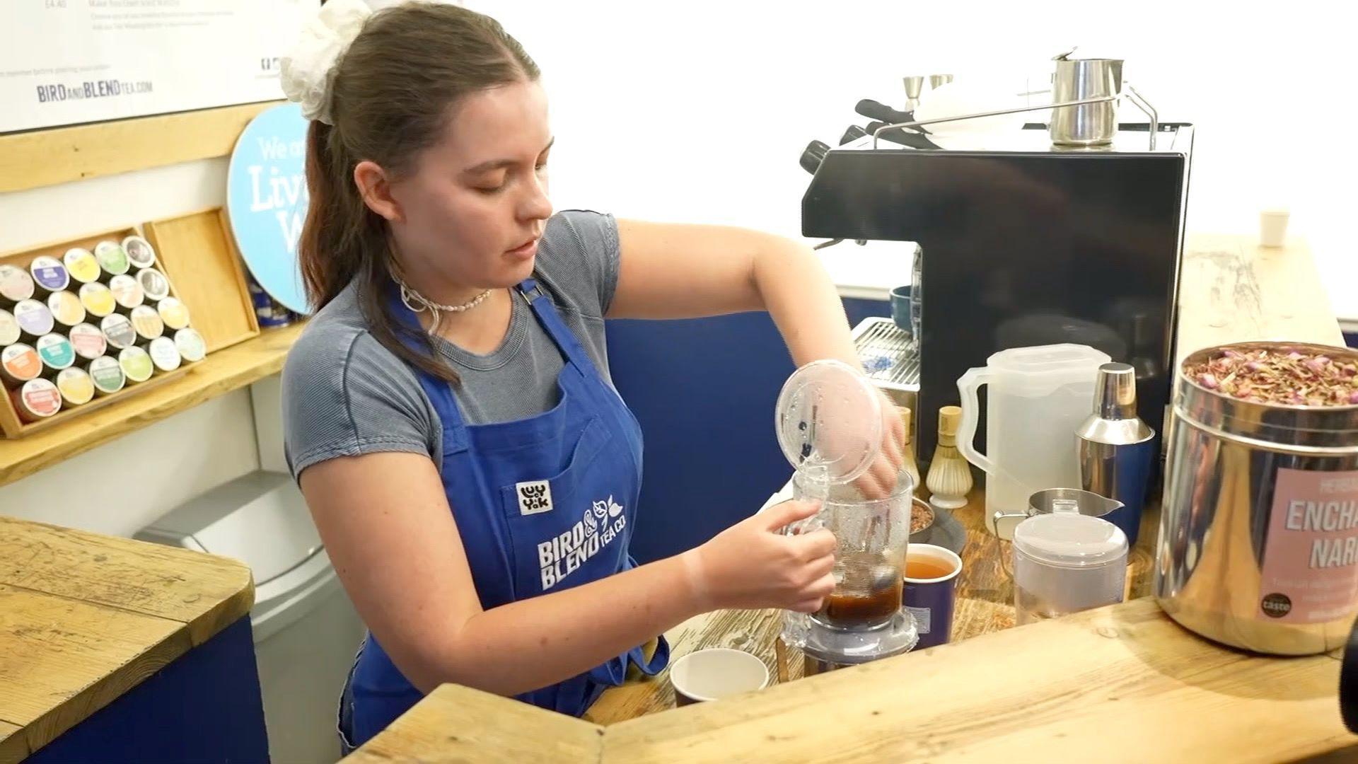 Hannah works at Bird & Blend and is wearing a blue apron with the logo on it. She is making coffee beside a coffee machine and there are coffee pods behind her. 