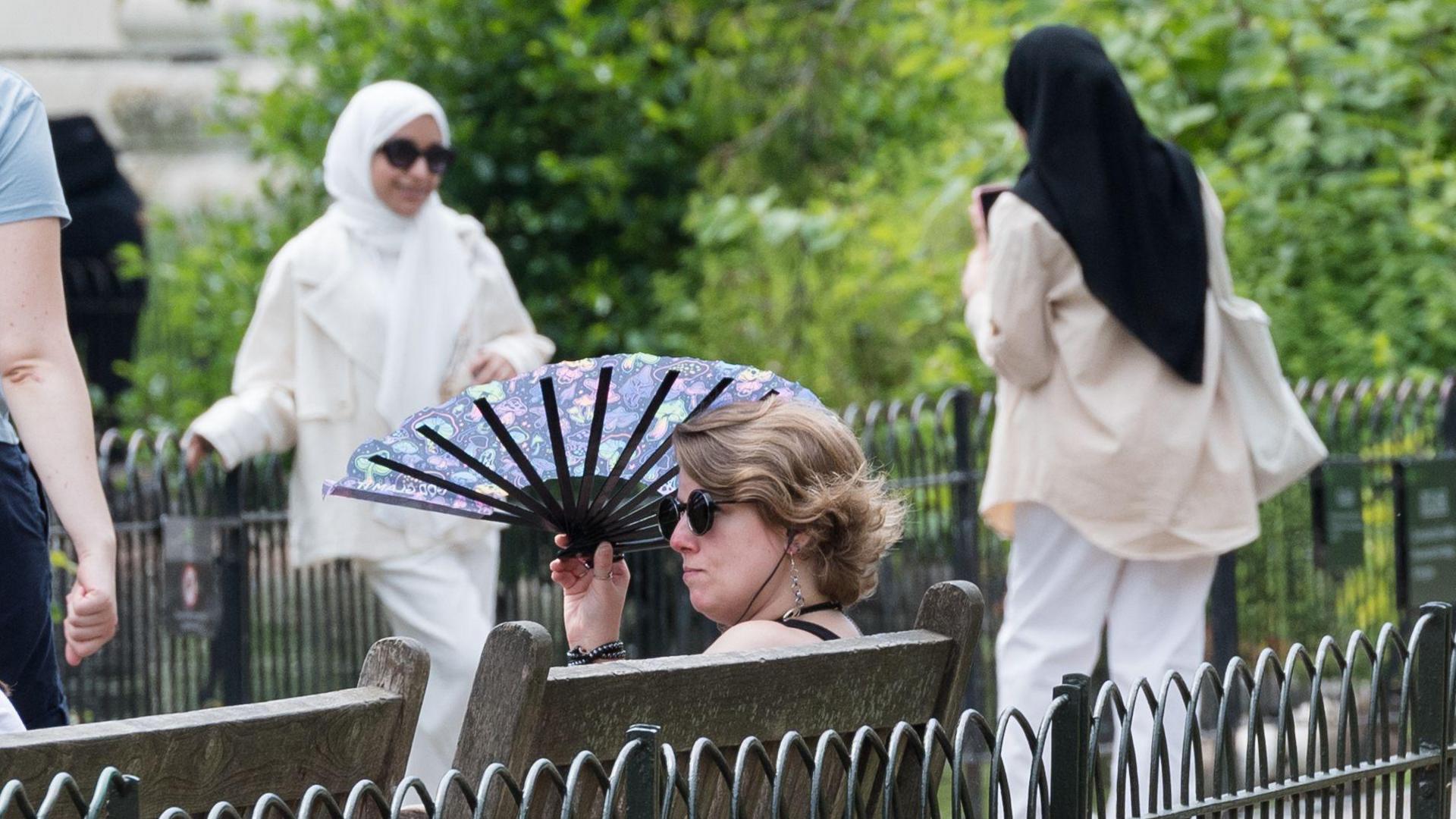 a woman shades herself with a fan while sitting on a bench as two women in the background takes pictures of one another