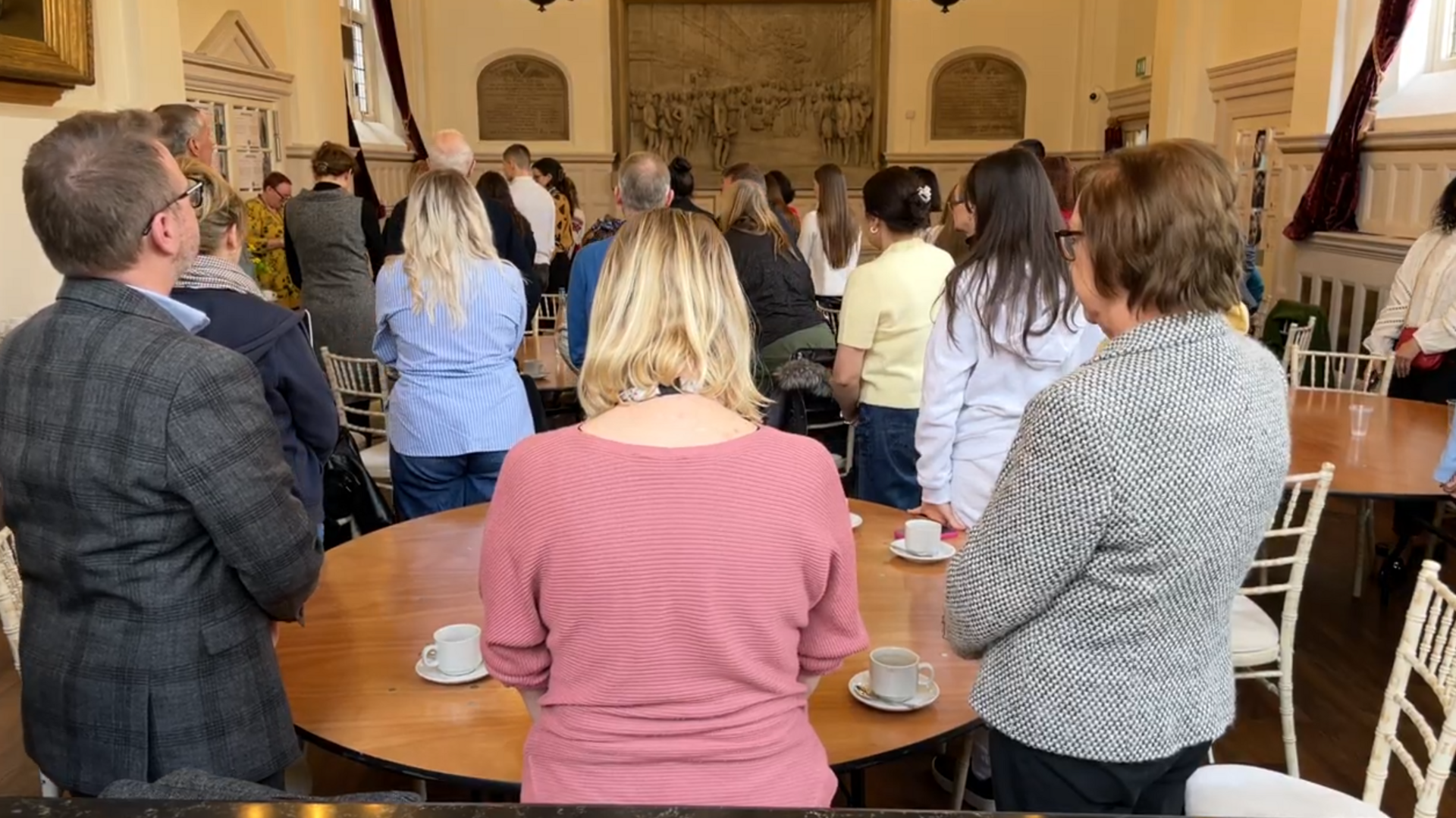 About 30 people standing around tables with their heads bowed as they take part in a minute's silence.