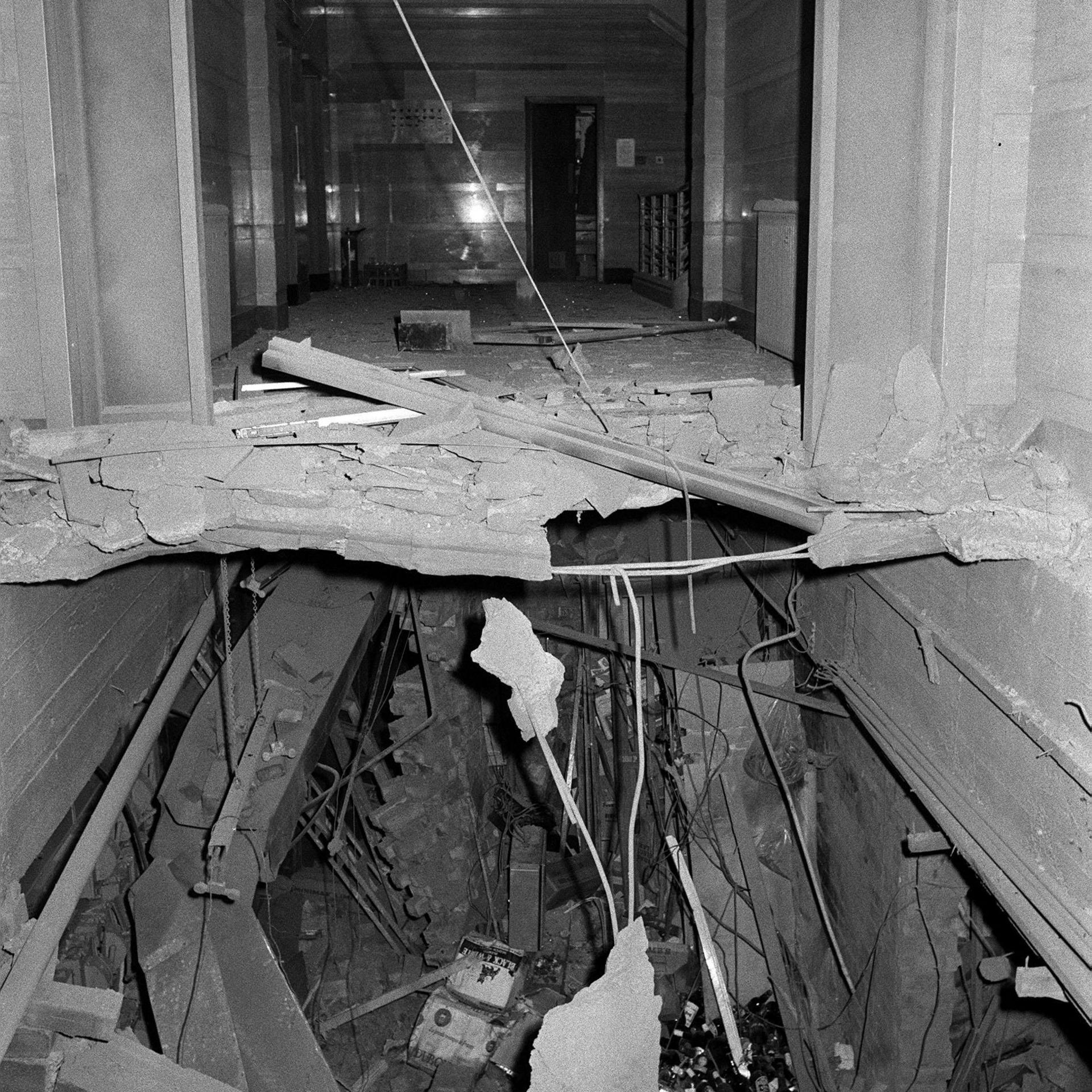 A black and white image showing a street-level ceiling blown away and a cavity into an underground room. Rafters and wiring hang down and rubble and bottles are strewn across the floor.