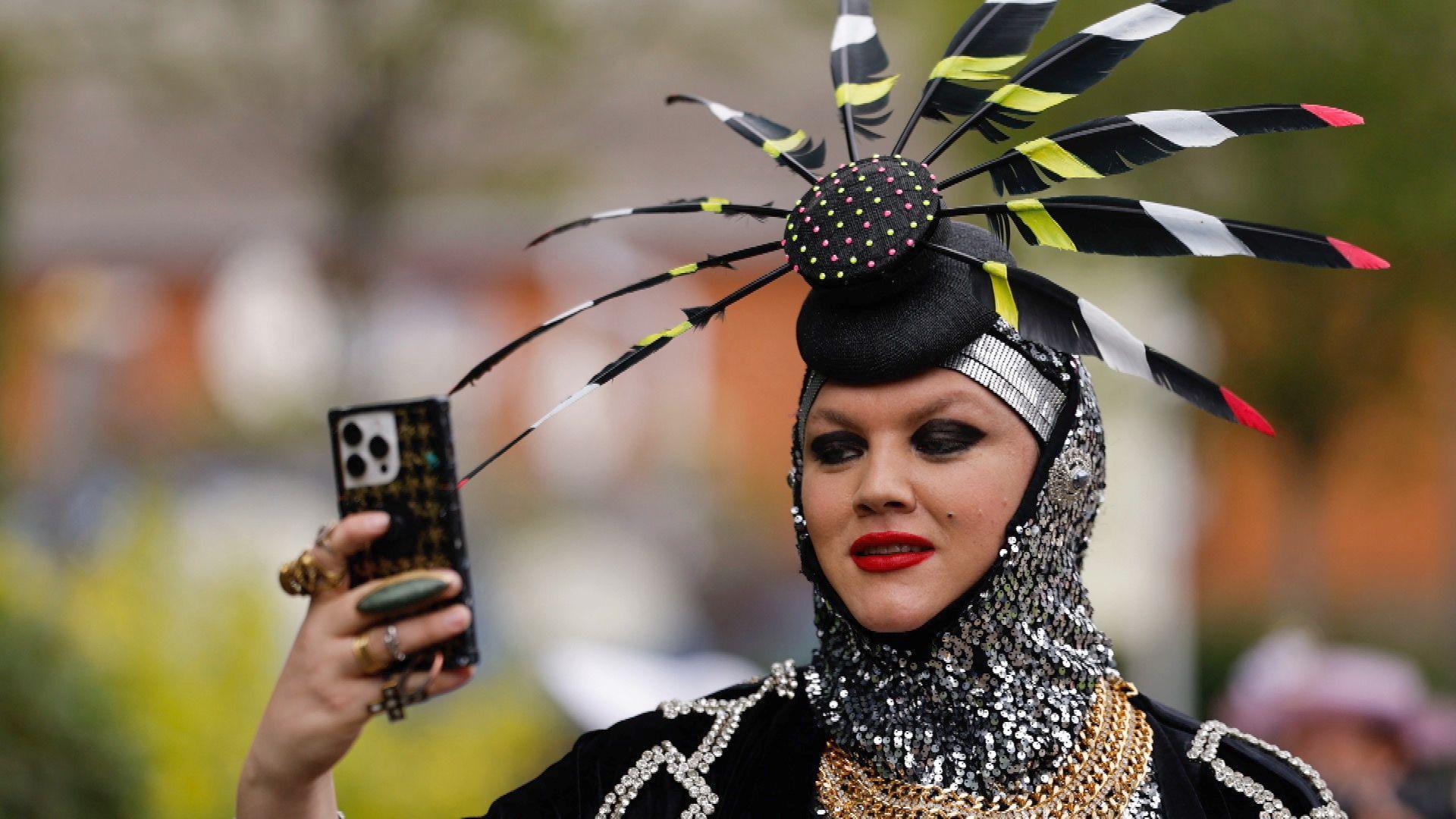 A woman is holding up her phone to look at the screen. She has a number of large ornate rungs on her right hand, which is holding the phone. She wears heavy black eyeshado and a bright red lipstick. On her head she wears a fascinator hat, which has a black base with a number of black, yellow, white and red striped feathers sticking out of it. She has a silver sequined hood over her head and a heavy gold chain around her neck.