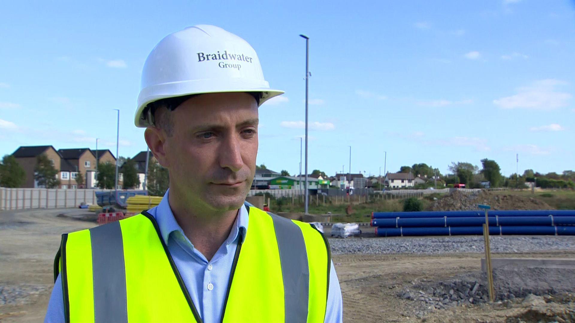 A man wearing a blue shirt underneath a yellow high-viz vest and a white hard hat with Braidwater group written on it, in the background is a building site 