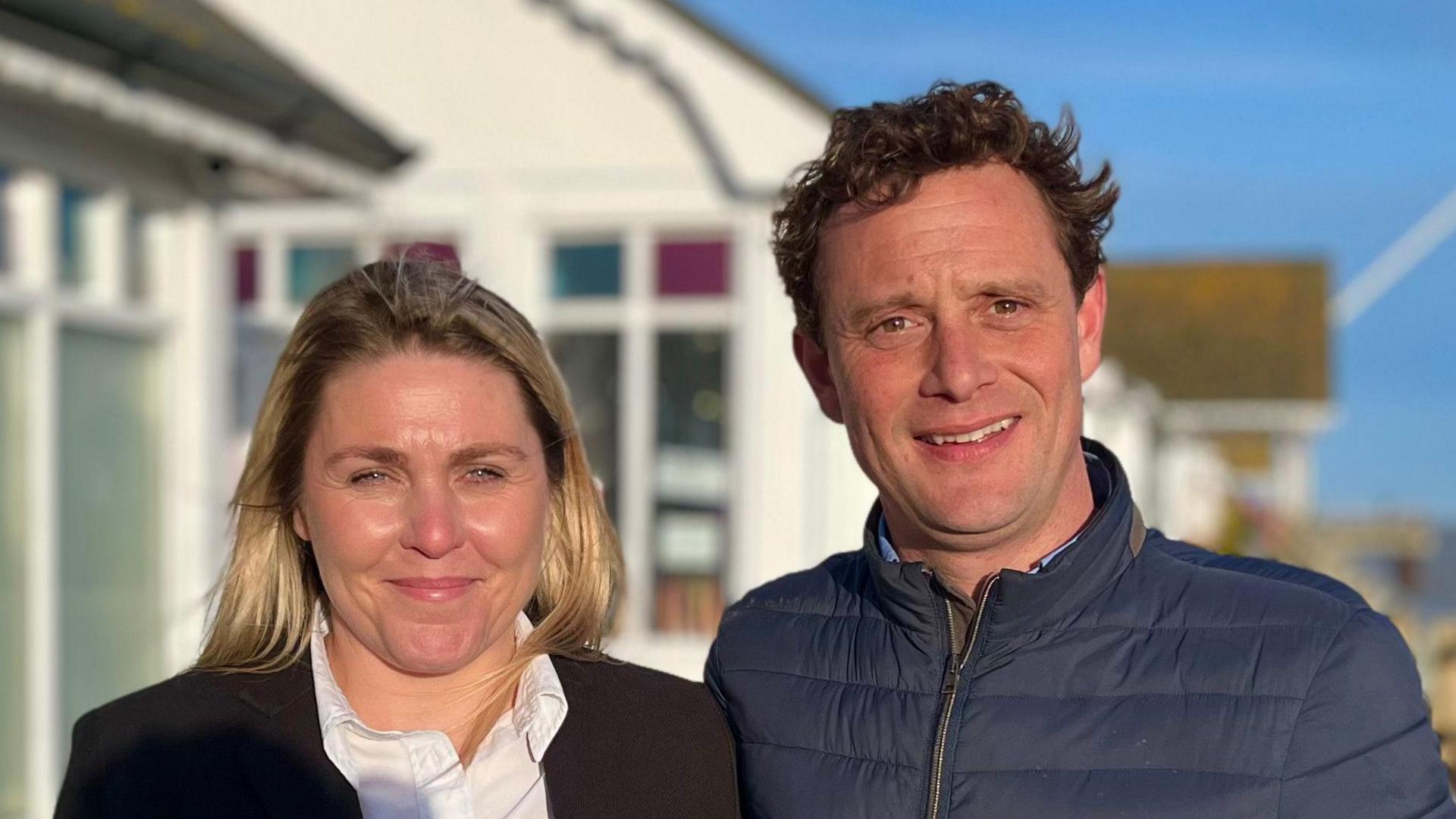 Amy and Charles Barwick smile at the camera on Southwold Pier. Amy has long blonde hair and wears a black blazer with a white shirt underneath. Charles has short brown hair and wears a blue puffer jacket. 