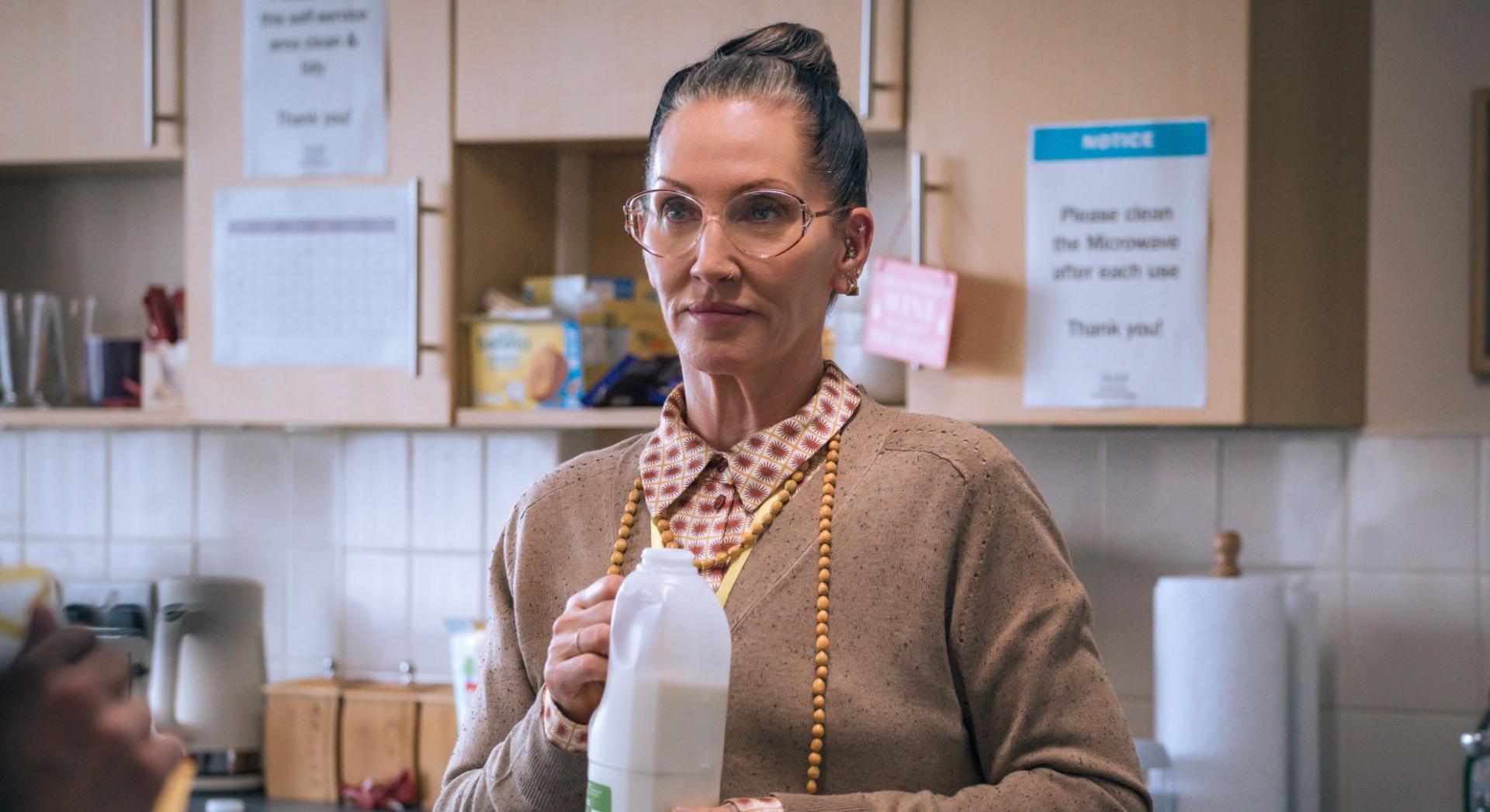 A woman with a slick bun and glasses hold a bottle of milk with the lid off. She wears a brown cardigan and wears a blouse with red dots. 