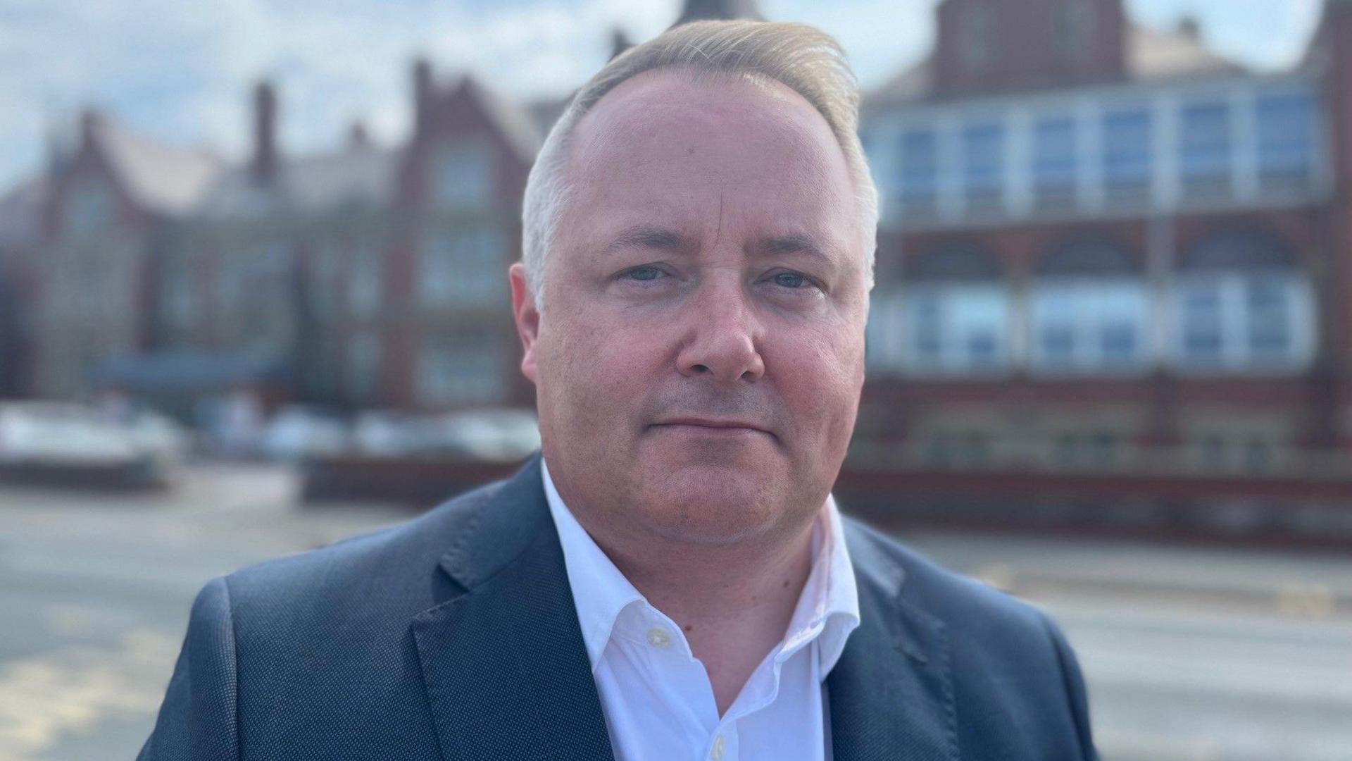 Darren Millar, standing in front of the Royal Alexandra Hospital in Rhyl. He is wearing a dark blue suit jacket with an open-necked white shirt. He has short blonde hair which is quiffed and it is a head and shoulders shot of him. 