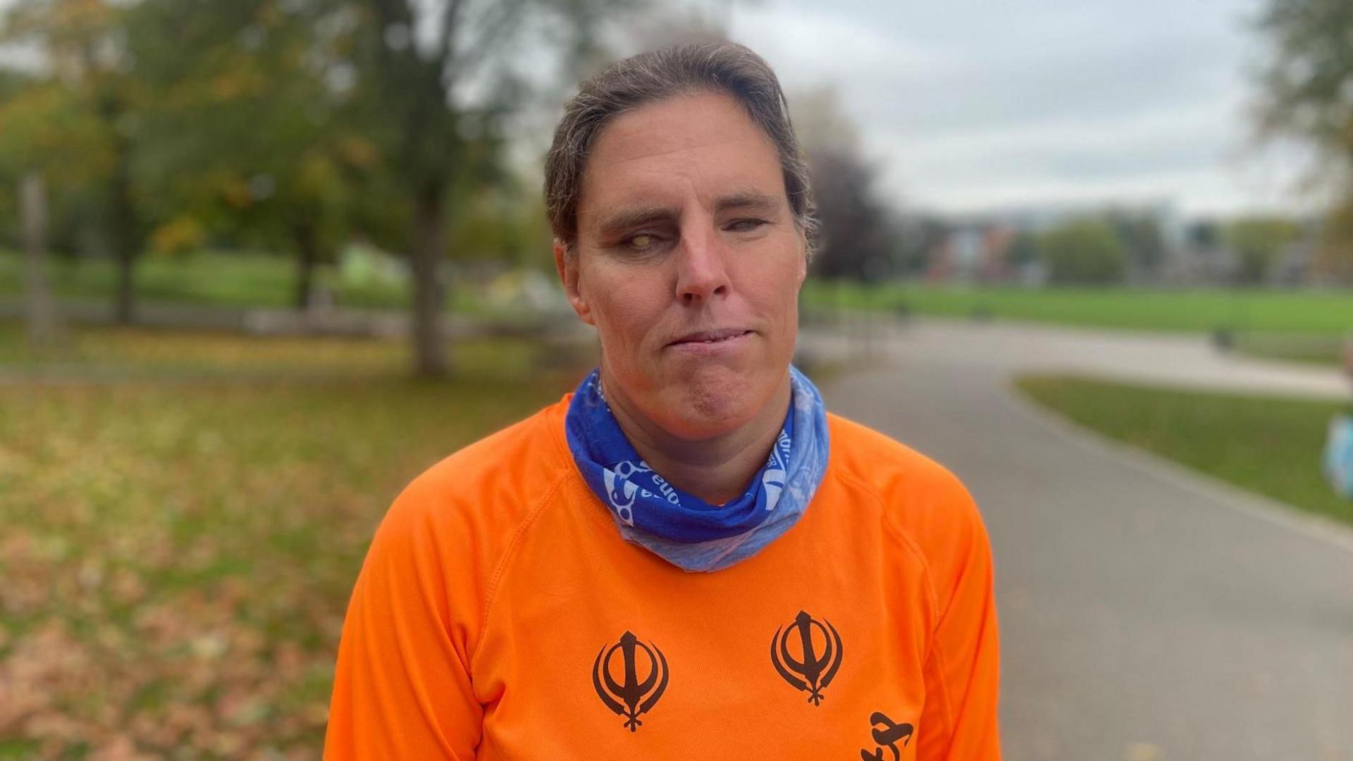A woman with dark hair tied back smiles shyly at the camera. She wears a bright orange running top and blue neckerchief