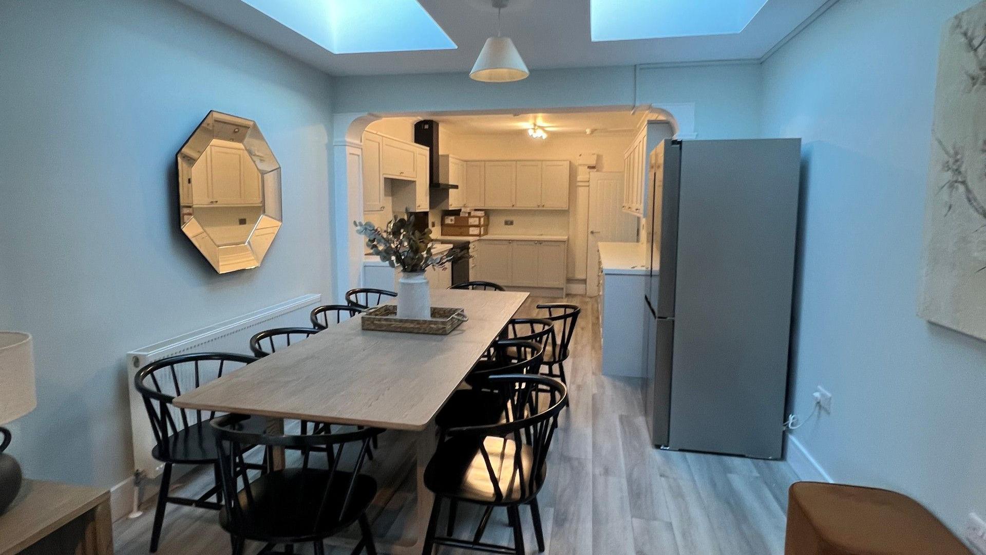 A modern kitchen-dining room. There is a large wooden dining table with six black chairs arranged around it. There is a large grey, free-standing double fridge-freezer against the wall to the right. A large ortagonal mirror hangs on the opposite wall. Beyond is a new kitchen with cream cupboards and a black cooker.