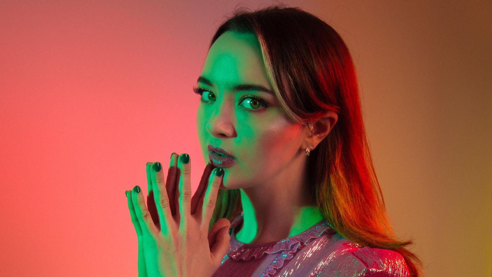 Fern Brady posing for a promotional picture, lit in red and green artistic light. She has long dark hair and green eyes. She is wearing a pink sparkly dress and is holding her hands in a prayer gesture under her chin. She is looking sideways at the camera.