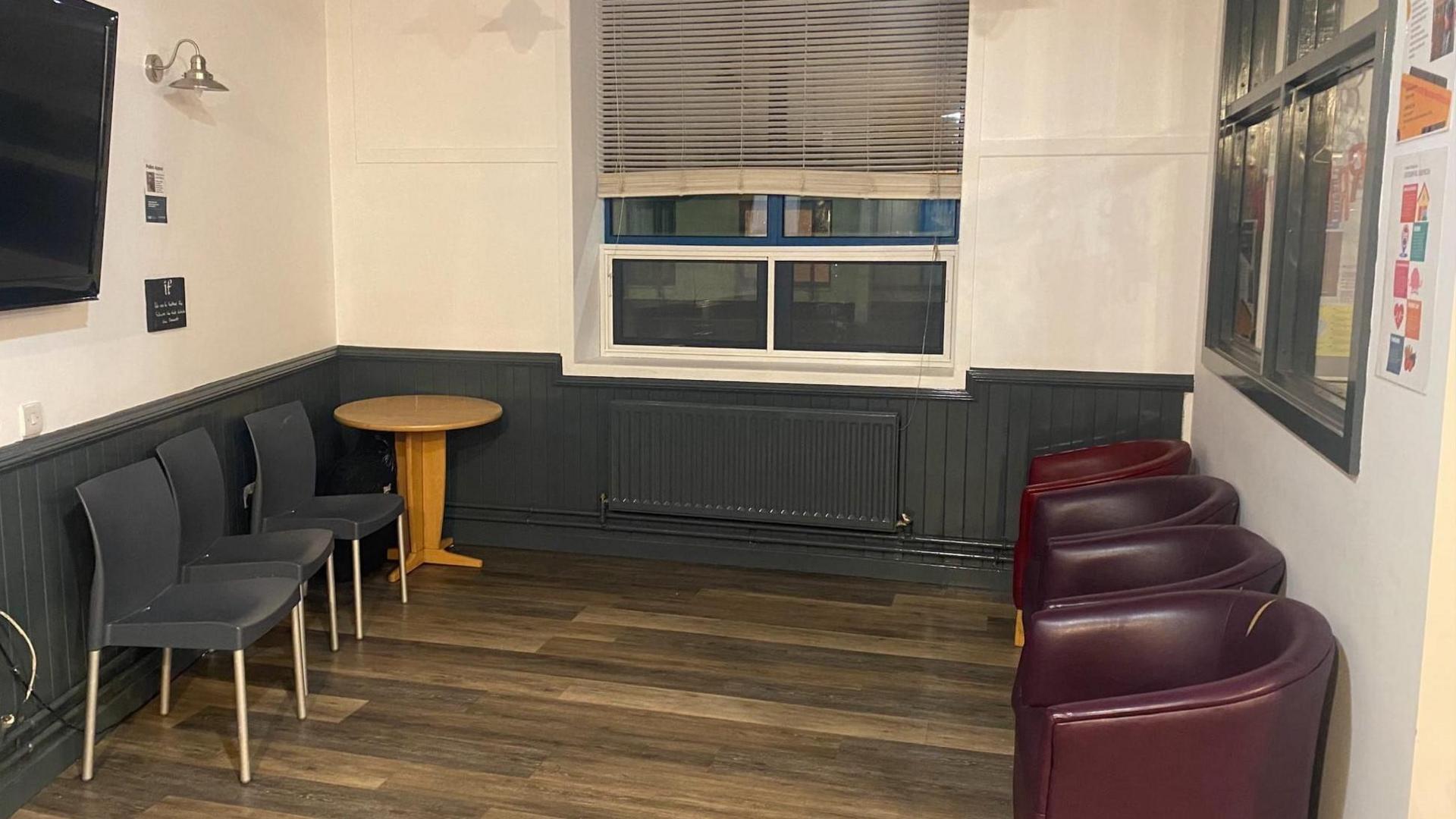 Four red leather seats lined up against the wall. Three grey chairs lined up against the wall. A little table sits in the corner. 