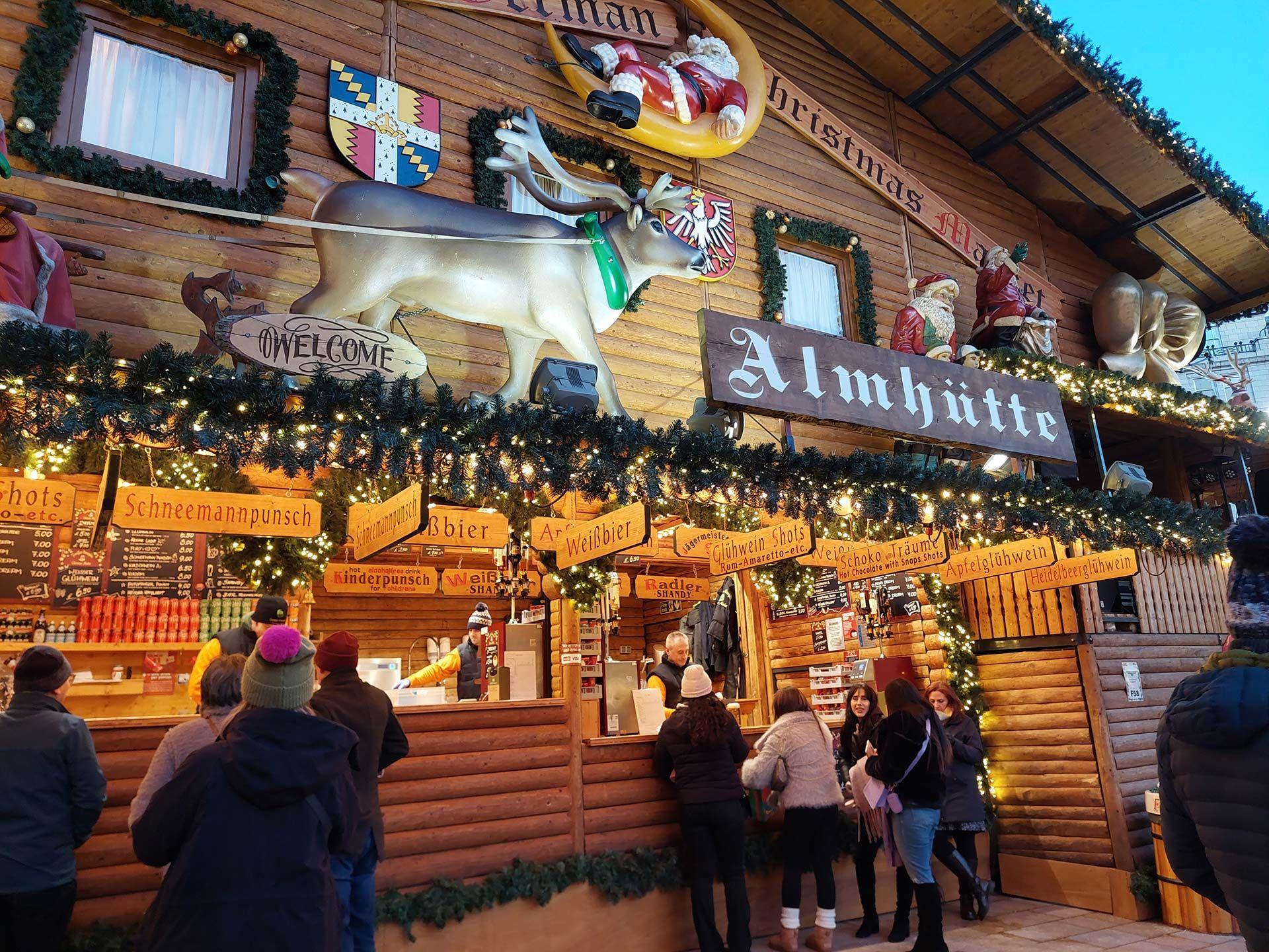 A stall selling alcohol at a Christmas market. The wooden stall is adorned with fake Christmas tree sprigs, golden fairylights, decorations including a giant reindeer and Santa ornaments, signs saying "Almhutte", "Weissbier", "Schneemannpunsch". A group of people in warm winter coats and woolly hats stand in front of the stall, being served by staff