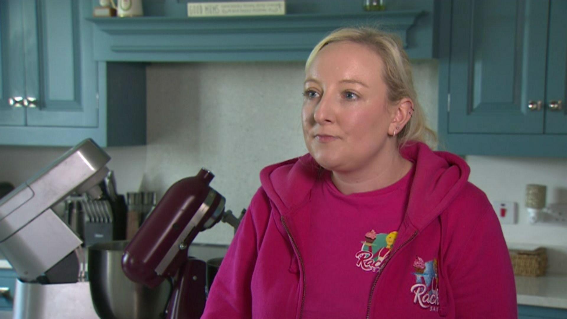 Rachel McGowan, with blonde hair and wearing a pink t-shirt and hoody, stands in her kitchen. There are two mixers and kitchen utensils behind her. The kitchen cupboards are blue.