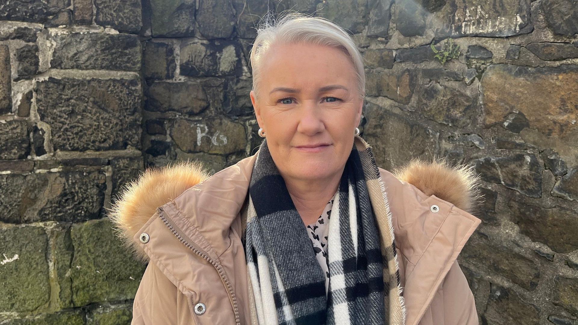 DUP councillor Tracy Kelly. A woman with short, blonde hair smiles at the camera. She is standing in front of a grey stone wall and is wearing a tan puffer rain coat and a black and white plaid scarf.