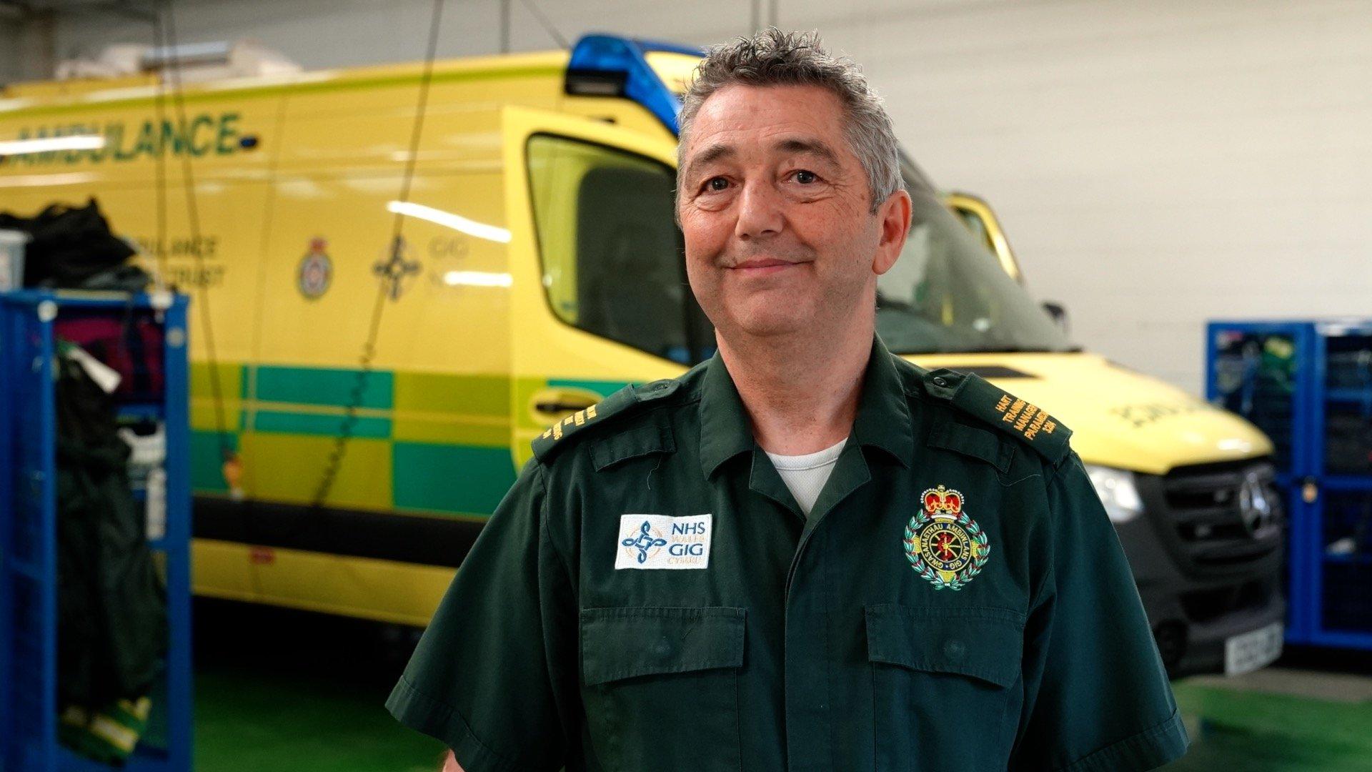 Giles has short, grey hair and is wearing a green uniform. He is stood in front of one of the large response trucks.