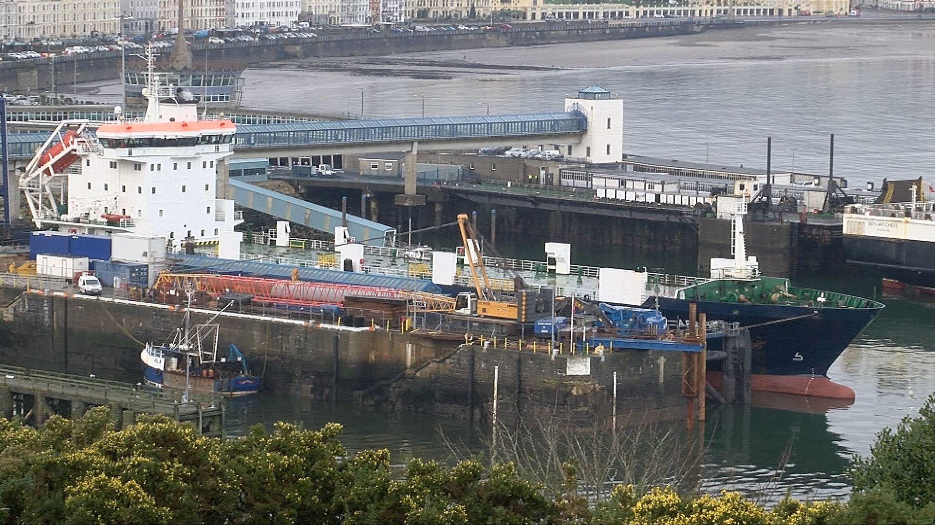 A long blue ship called the MV Arrow in the harbour with a white cabin and a green top. The ship is next to a long jetty and has a blue bridge behind it. 