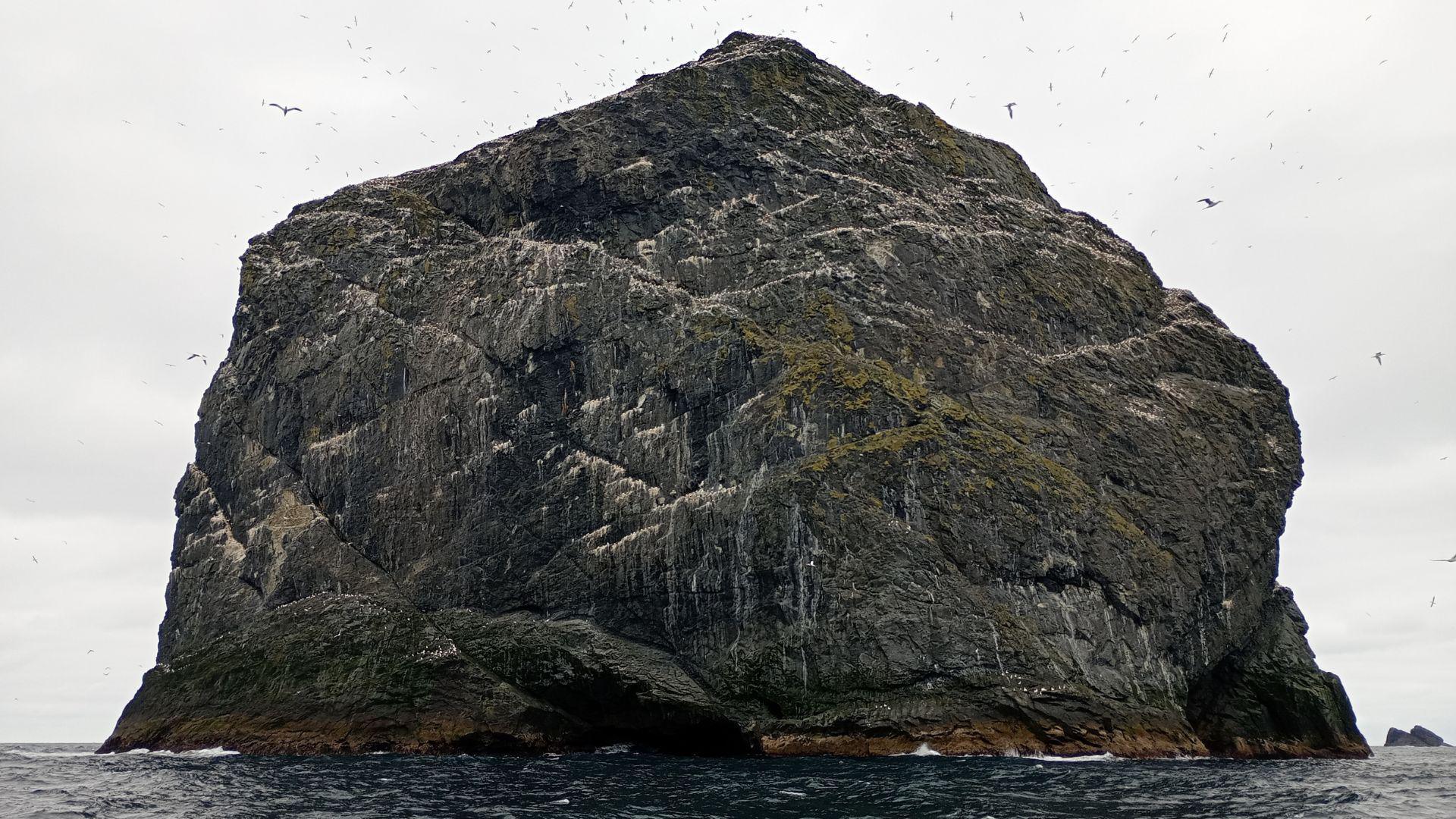 A dark island on an overcast day. The island is mainly made up of dark colours with a few streaks of white on the rocks. The sea is in the foreground and is also dark. The sky overhead is grey.