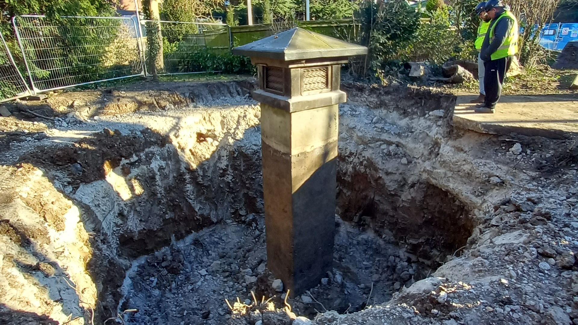 A dug-up area revealing a deep excavation with a WWII-era ventilation shaft emerging from the ground, surrounded by construction fencing and workers in high-visibility vests inspecting the site.