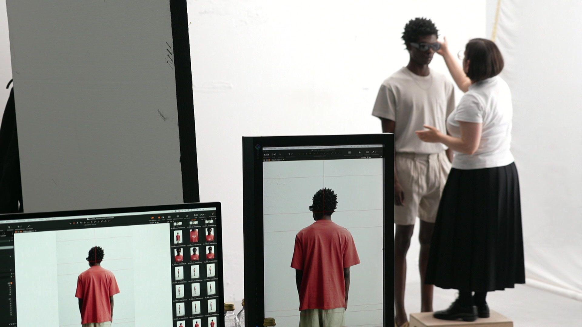 Male model standing wearing a light-coloured T-shirt and shorts with a woman standing on a block in front of him adjusting his sunglasses. Computer screens in the foreground show pictures of the same model taken from behind with him wearing a red T-shirt.