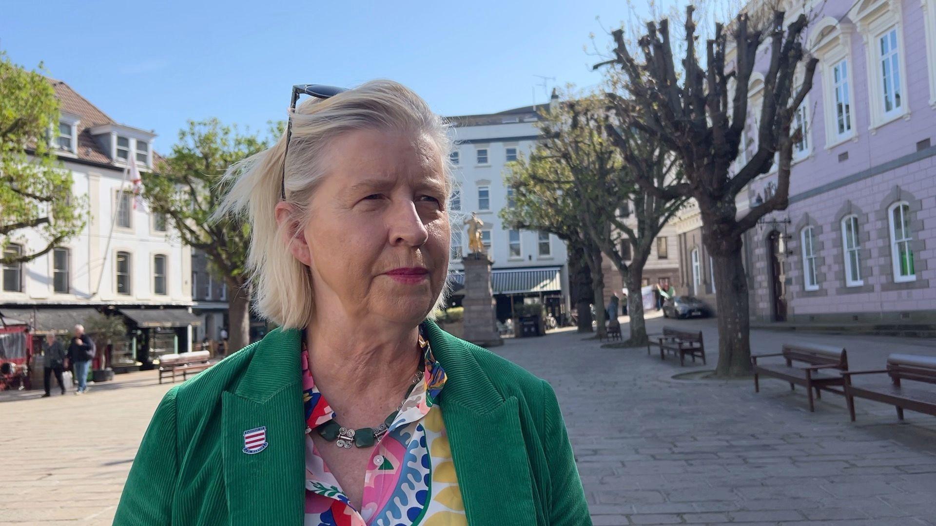Constable Karen Shenton-Stone is wearing a green blazer and a yellow, pink and blue shirt. She has short blonde hair and is stood in Jersey's Royal Square.