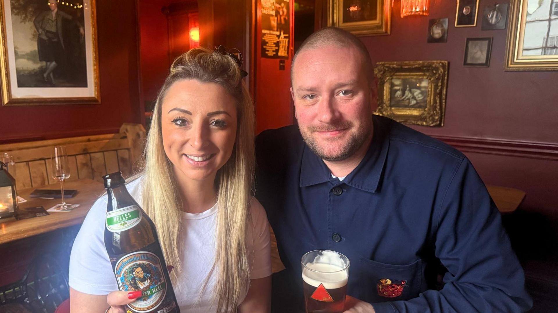 Two people sit together in a warmly lit pub with deep red walls and framed artwork. The woman on the left holds a bottle of beer, while the man on the right has a pint glass of amber ale. They’re seated at a wooden table with red cushioned stools, surrounded by string lights and other bottles and glasses.