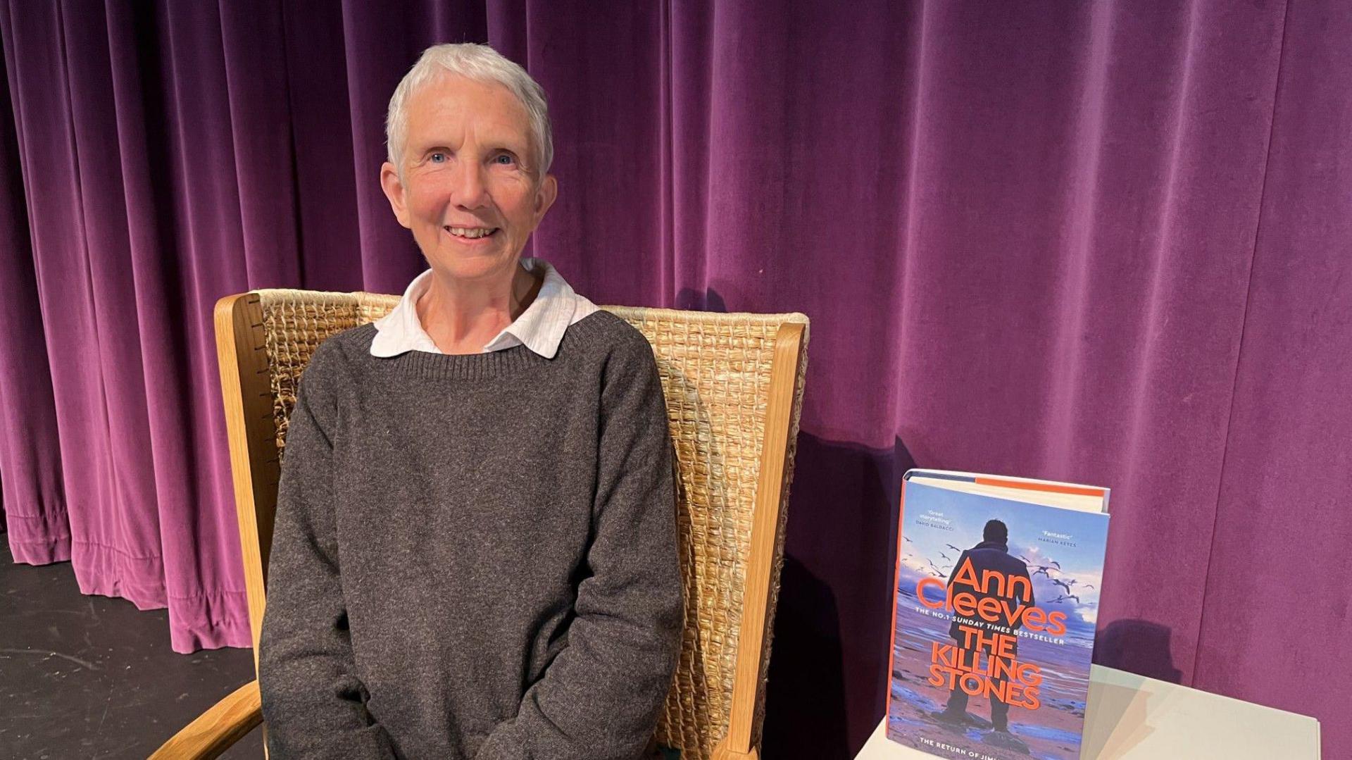 Author Ann Cleeves, who has short white hair sits smiling in a woven wooden chair. She is wearing a dark grey sweater over a white collared shirt. Behind her is a stage backdrop of deep purple curtains. Next to her is her new book, The Killing Stones which is standing on a small white table.