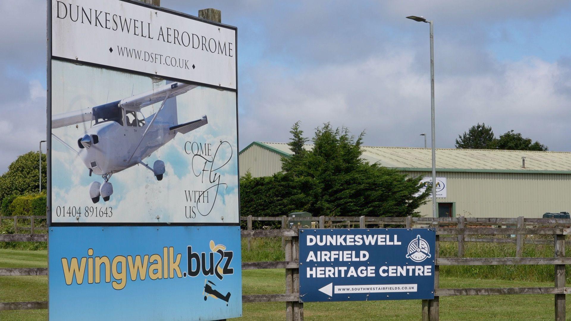 A sign showing a light aircraft with Dunkeswell Aerodrome written above. There is also a navy blue sign mounted to a wooden fence that reads Dunkeswell Airfield Heritage Centre.