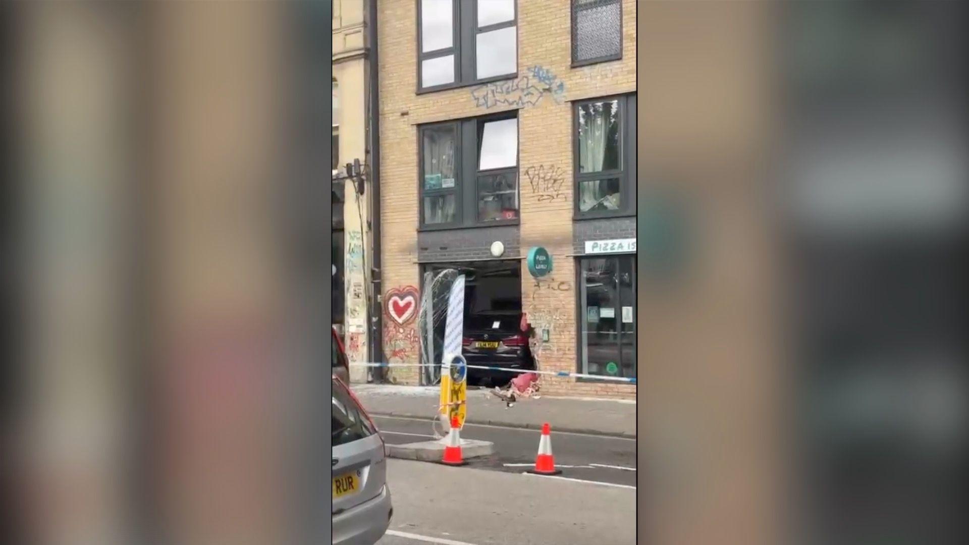 A black BMW that has crashed into a pizza restaurant. There is police tape and traffic cones cordoning off the scene.