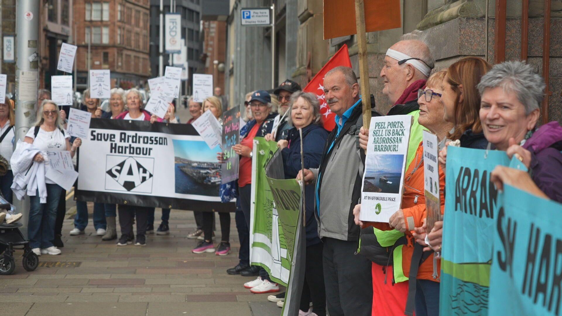 People holding banners and placards line the pavement of a Glasgow street. The visible largest banner reads "Save Ardrossan Harbour".