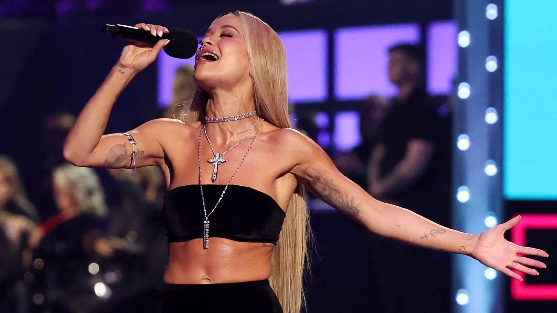 Rita Ora hosts the 2024 MTV Europe Music Awards at Manchester Co-op Live in Manchester earlier this month. She is singing into the microphone and has long blonde hair worn down. She is wearing a black boob tube and silver jewellery.