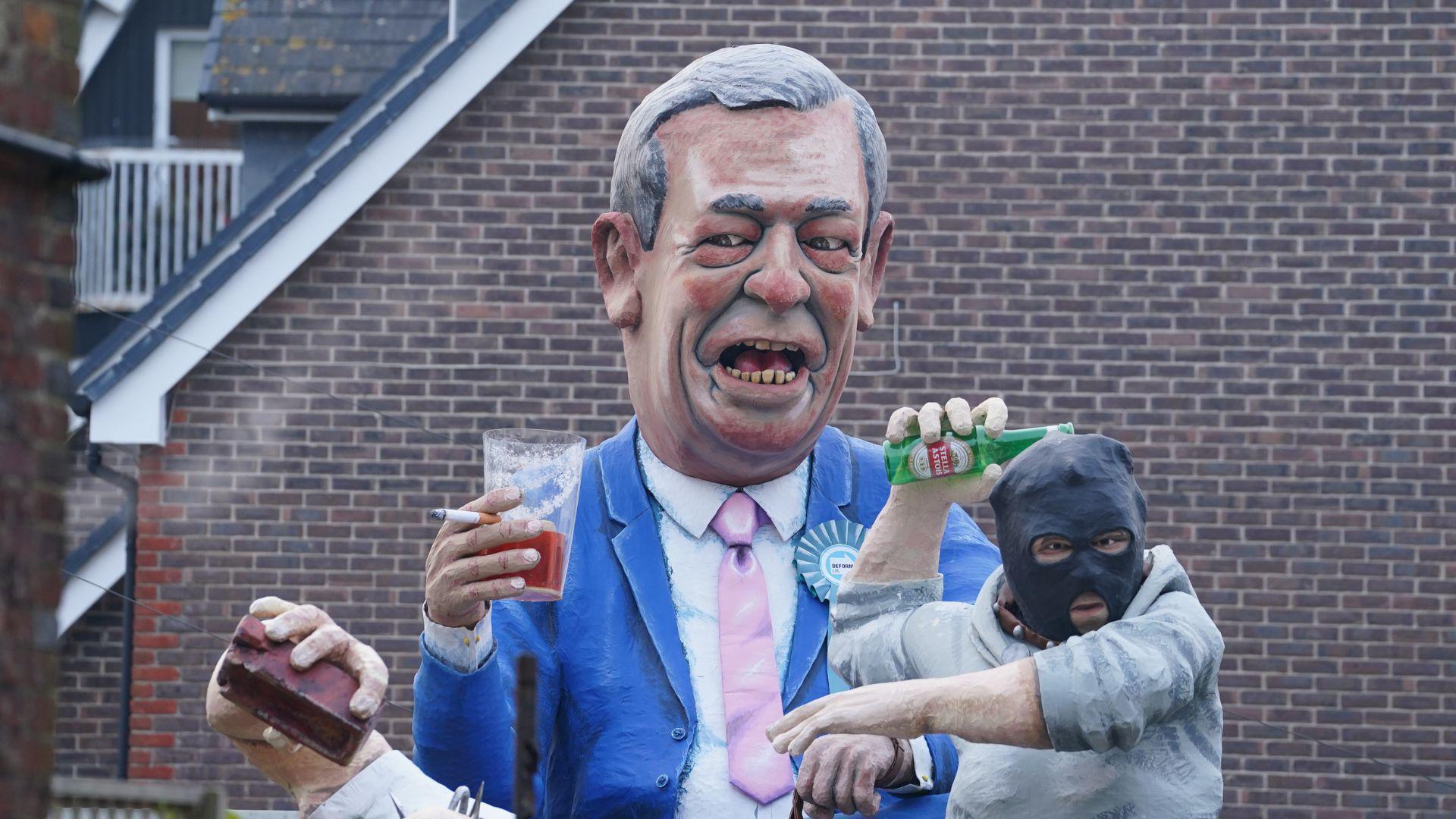 A large model of Nigel Farage holding a cigarette and a pint of beer. He is wearing a blue suit and pint tie. Next to him is a model of a person in a grey jumper wearing a black hood, throwing an empty glass bottle of Stella Artois