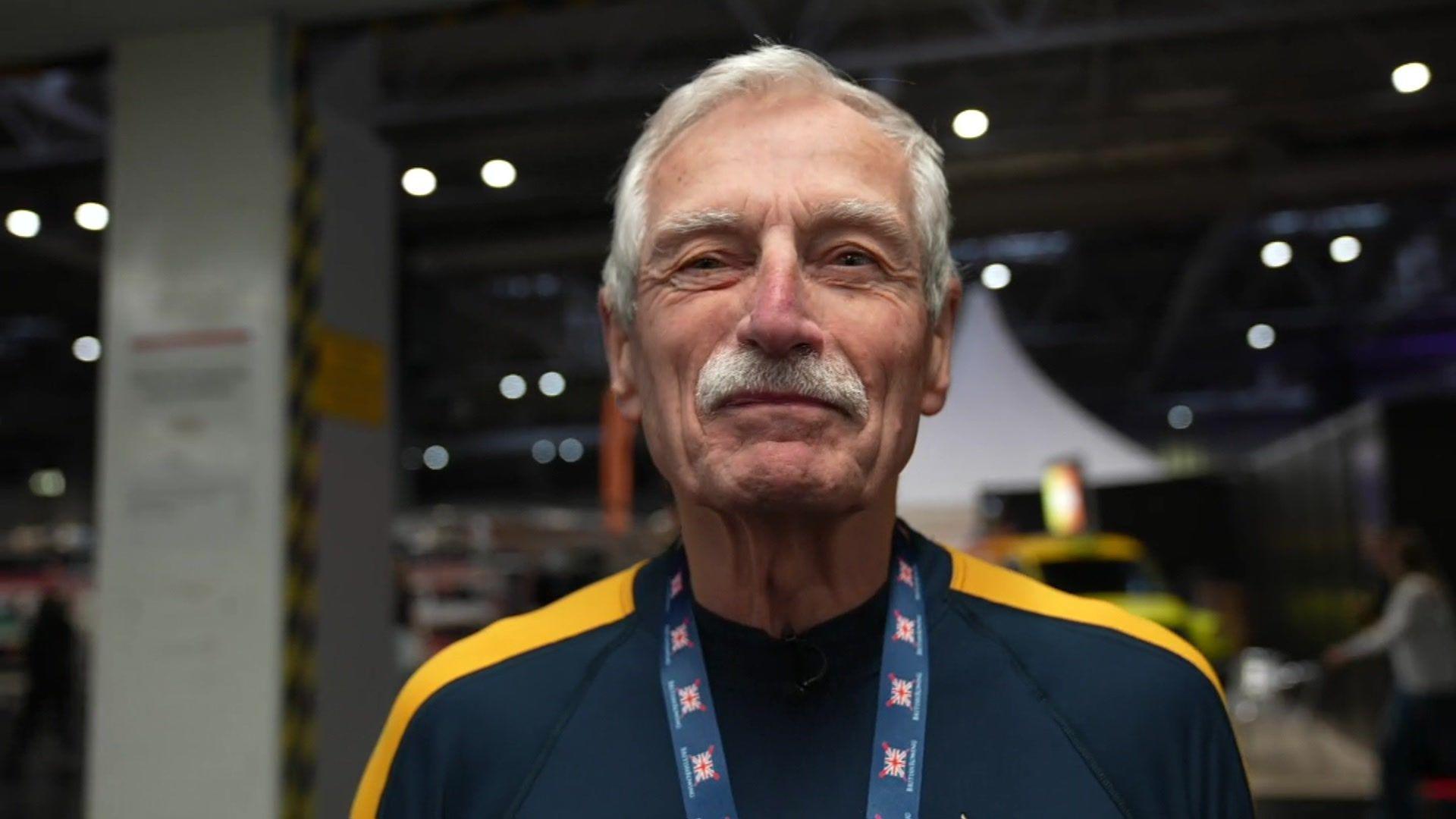 Roger Stainforth has a grey hair and a moustache. He is standing in a gym and is wearing his medal around his neck. 