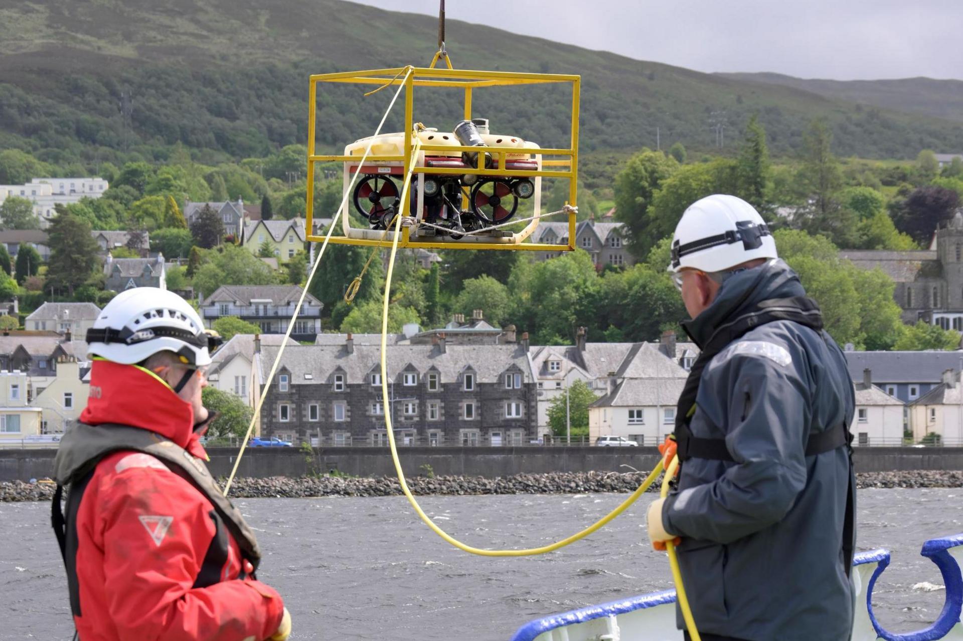 A small ROV is lowered into Loch Linnhe at Fort William by two men. The men are wearing overalls and white hard hats.