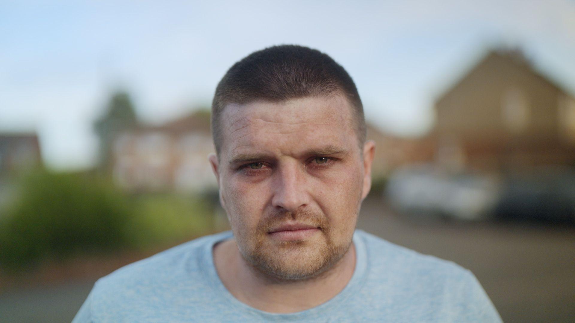 Ross Hart, a man with close-cropped brown hair and stubble, wearing a grey T-shirt, looking directly into the camera in a close-up shot with a shallow depth of field, so that the houses behind him are blurred