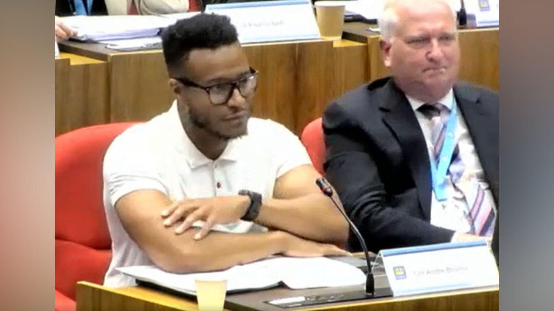 A screen shot of a council meeting - a councillor sits at a desk and is wearing a white T-shirt and glasses sits with his arms folded over some paperwork. There is a sign in front of him that reads Cllr Andre Bourne. Beside sits another man in a dark suit, white shirts and striped tie. There are other people sat at similar desks in the background. 