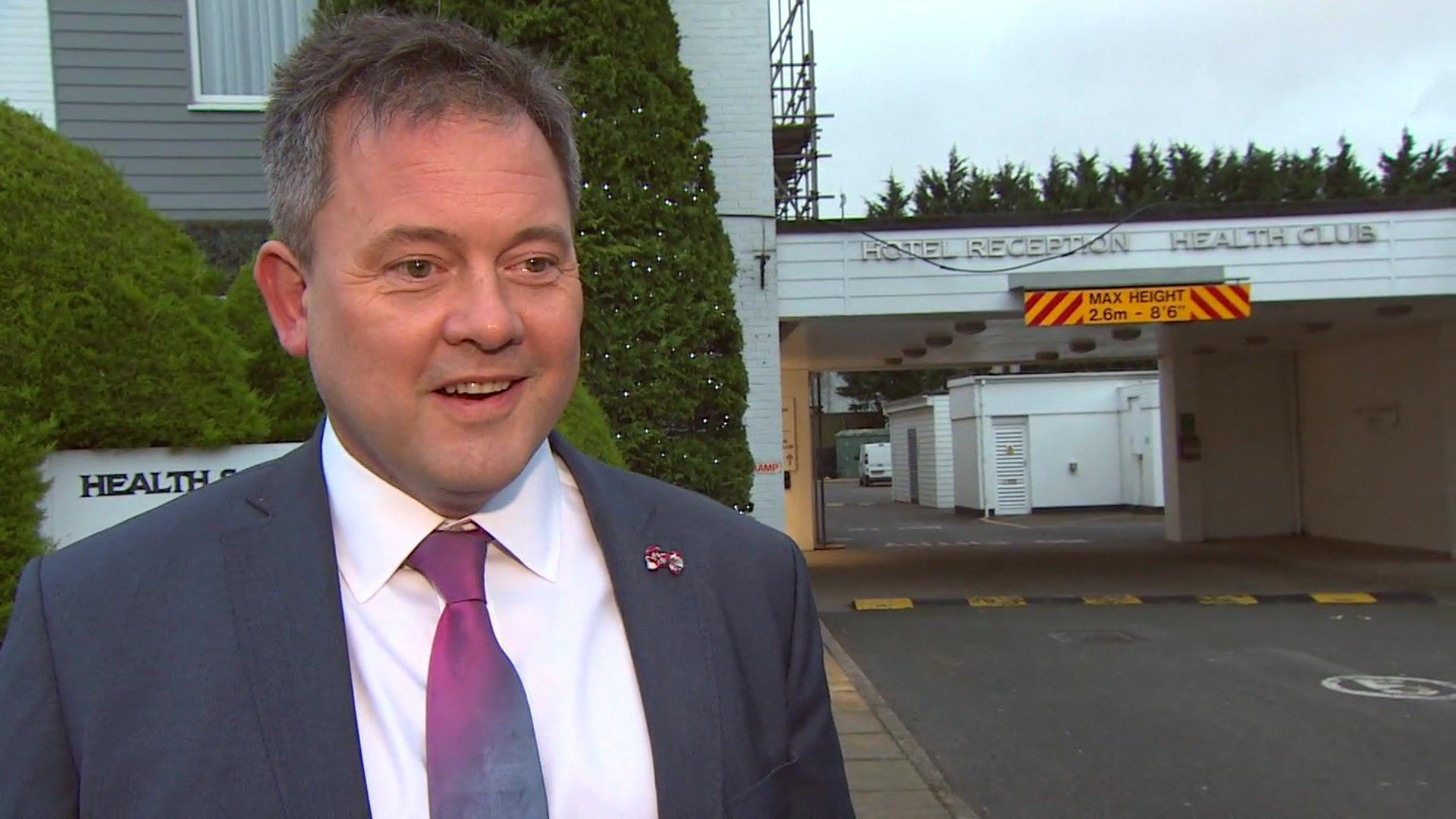 Close up of Peter Brend. He is standing outside of the hotel in Barnstaple. He is wearing a blaze, white shirt and a purple tie. Mr Brend is a white man and has dark grey hair.