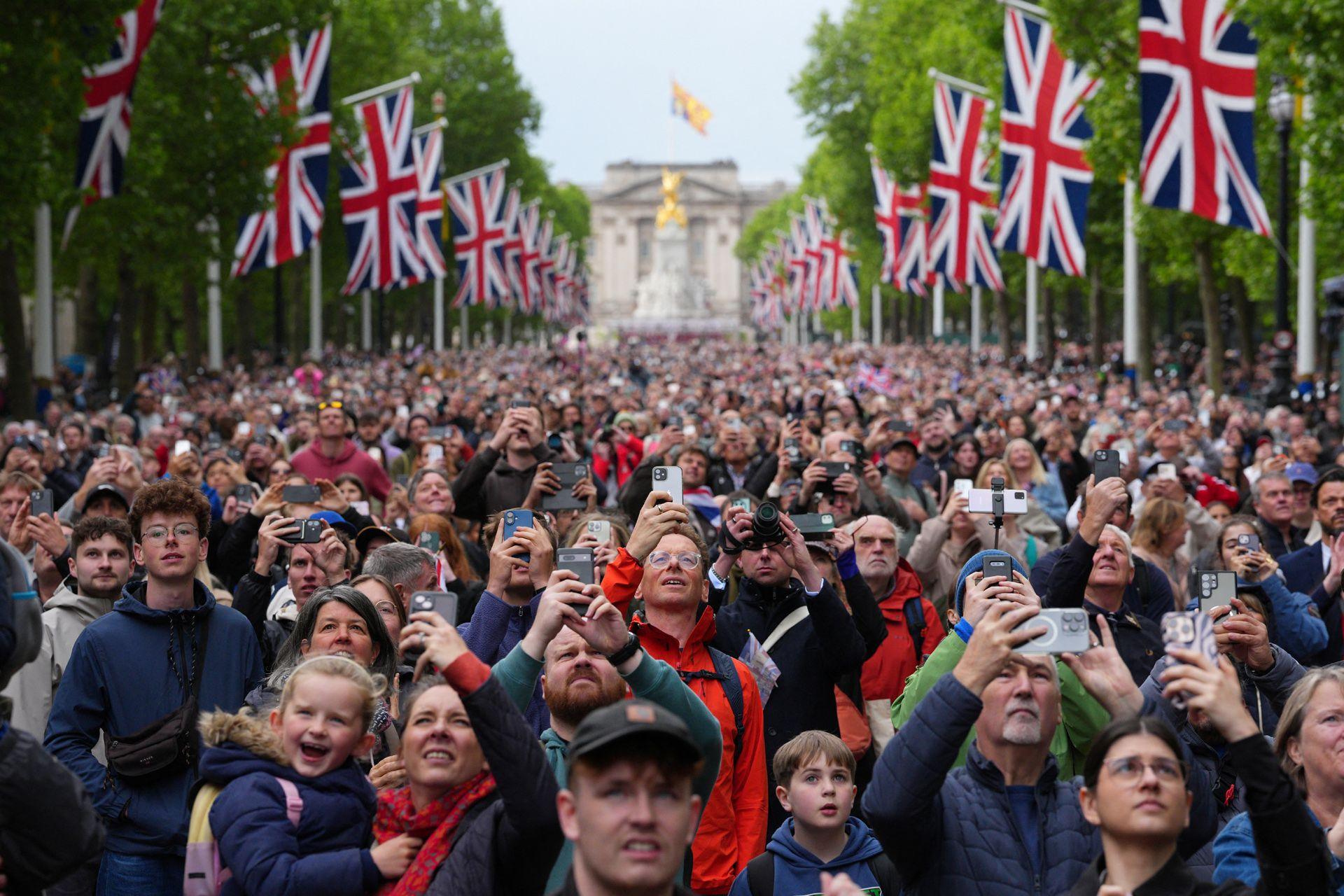 A huge crowd gather in the Mall after the procession, and look up to the sky while filming and taking photos with mobile phones, with union flags and green foliage lining the street and Buckingham Palace in the background.