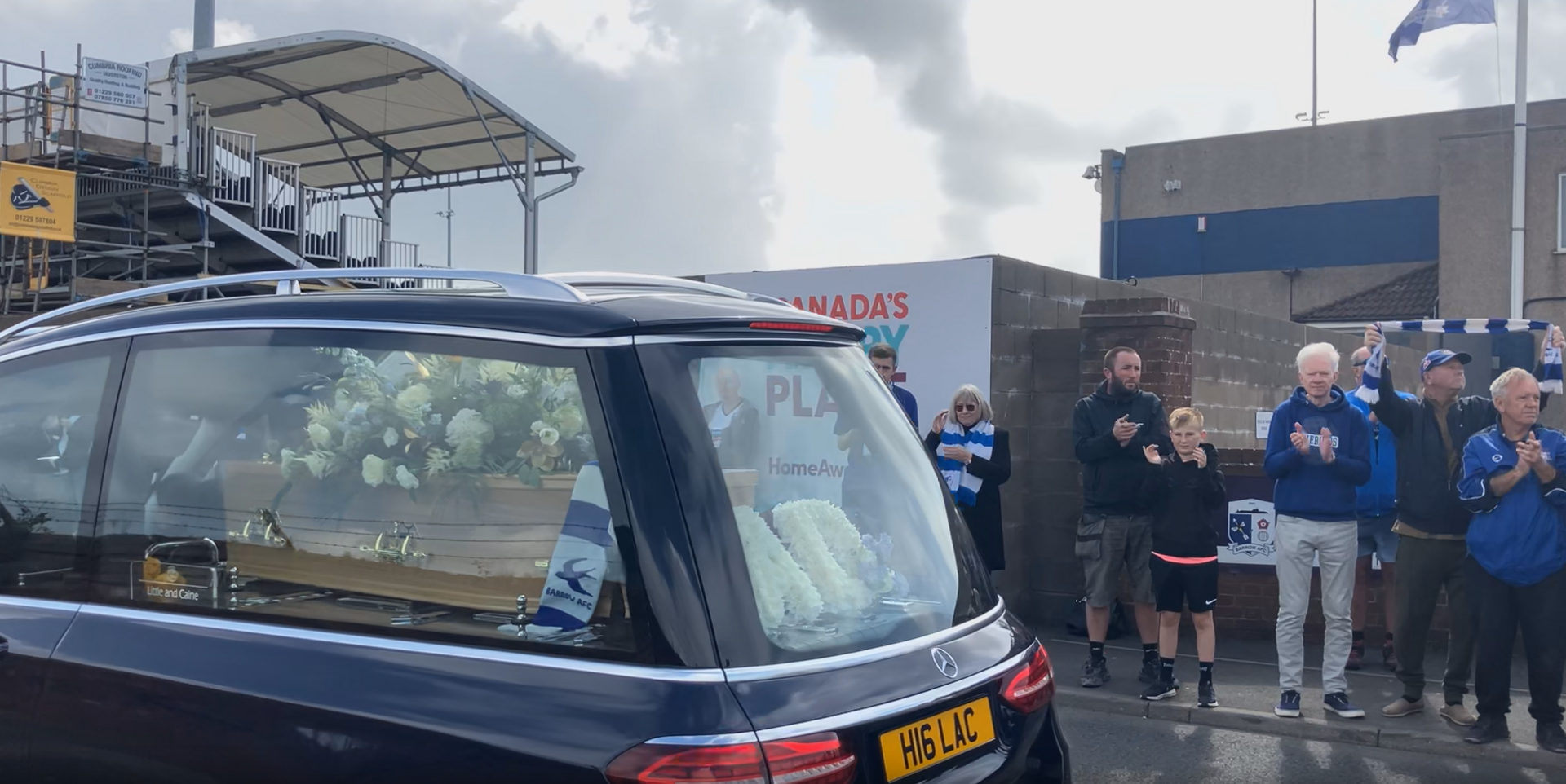 A hearse - with Mr Keen's coffin and flowers inside - drives along a street. Fans are clapping and wearing blue and white striped scarves.