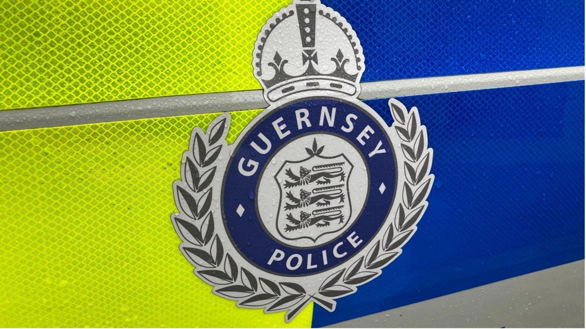 There is the side of a police car within the picture. At the centre of the screen there is the guernsey police logo. It is silver and blue colours.