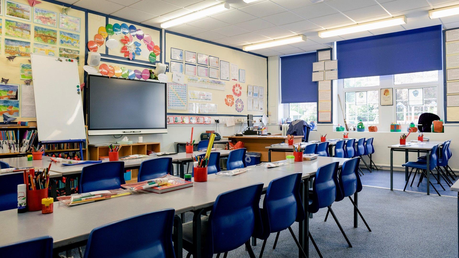 A bright, empty classroom with desks, chairs and pencils in the foreground