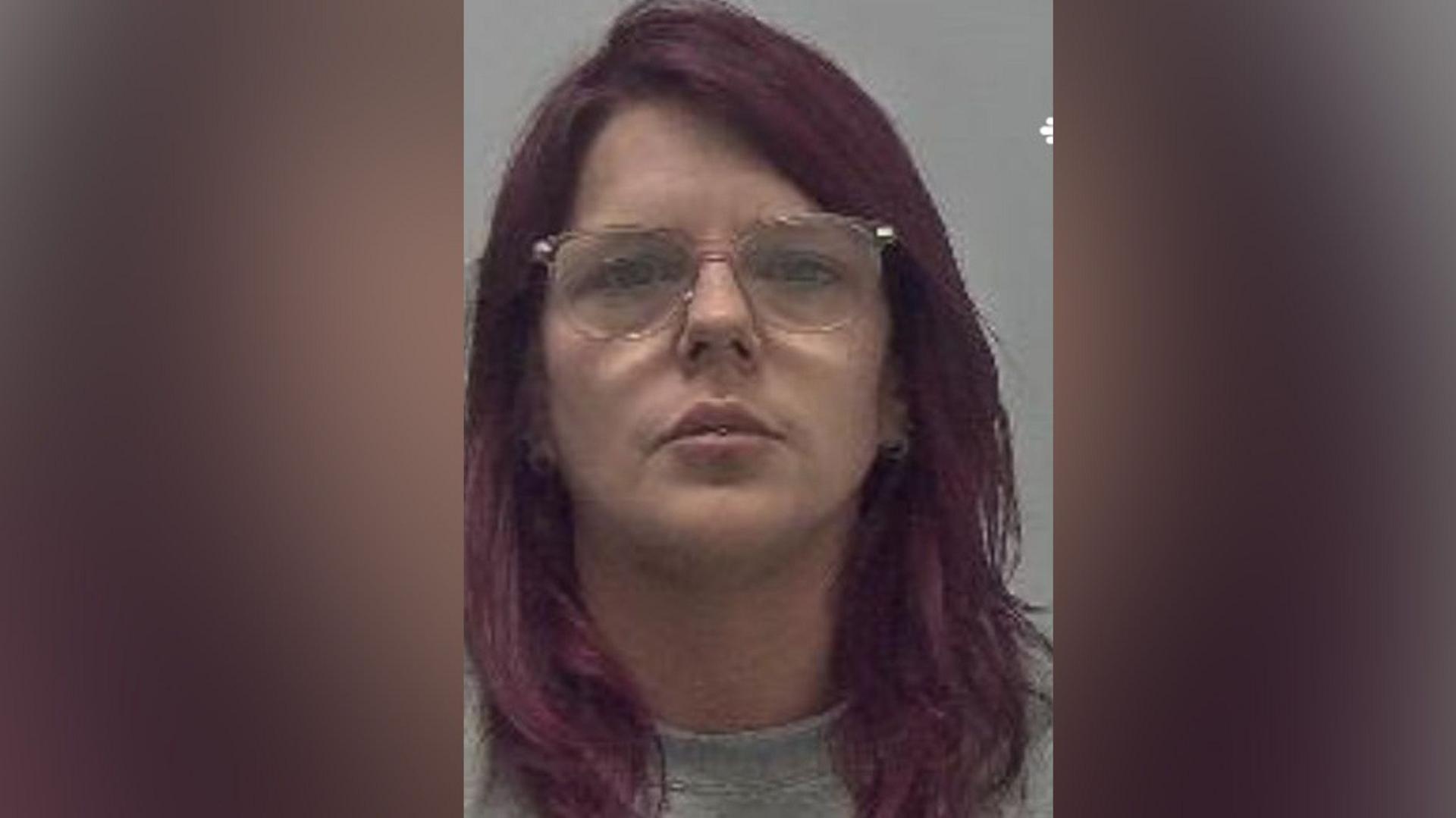 A woman with red hair and glasses wearing a grey top against a grey background