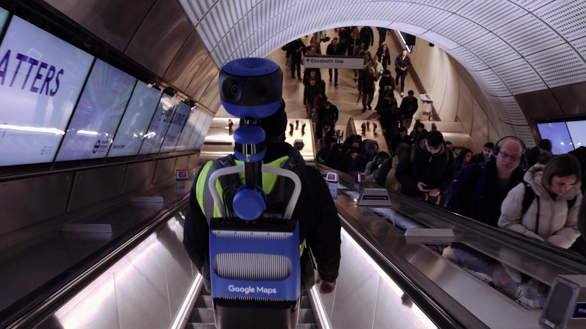 A person on the down escalator wearing a Google Maps filming apparatus of a blue rucksack style back pack with a camera poking out on a pole above the bearer's head. Commuters on the opposite escalator look askance at it.