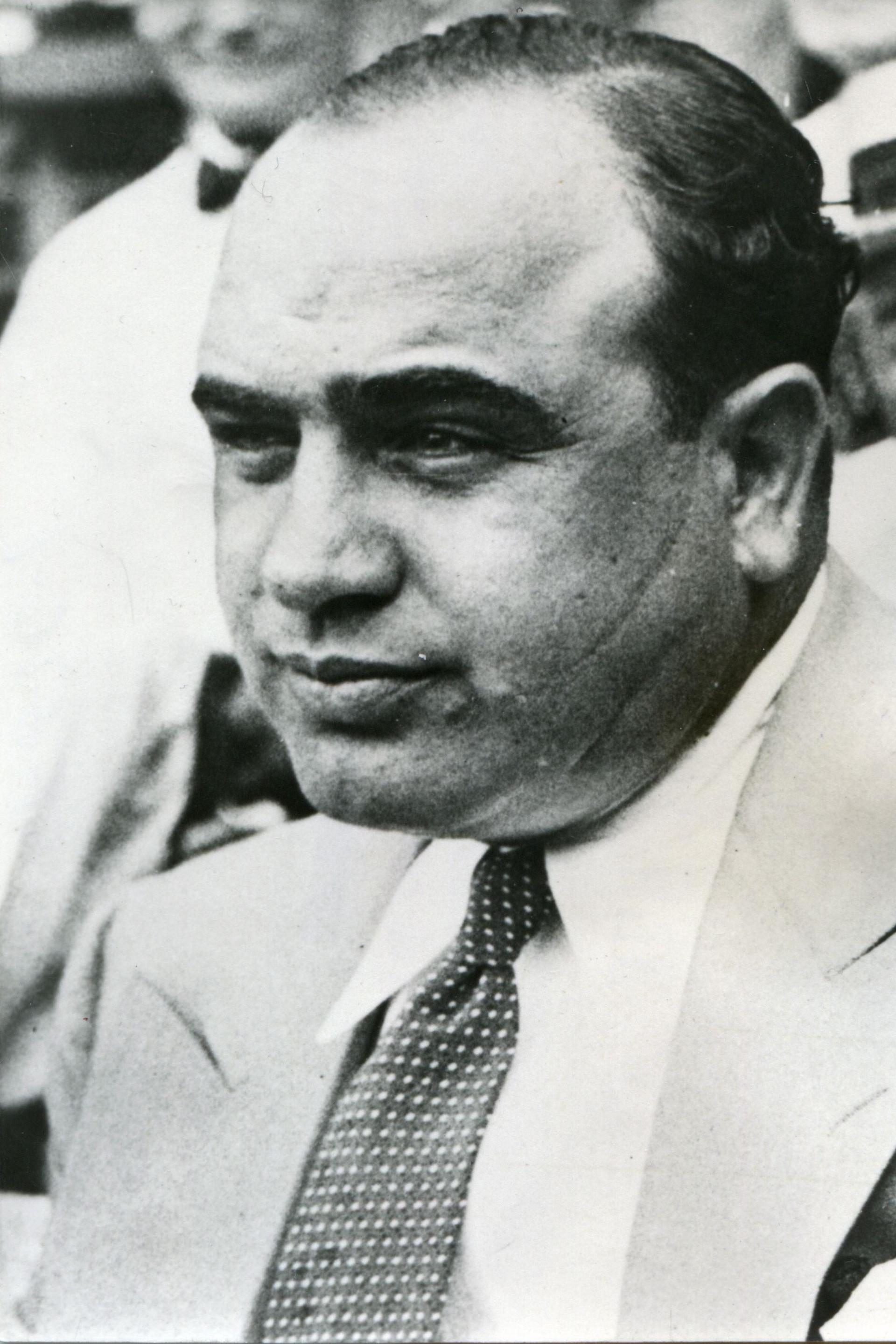 A balding Al Capone wearing a light coloured suit. He is smiling into the distance and his trademark scar is visible on his left cheek 