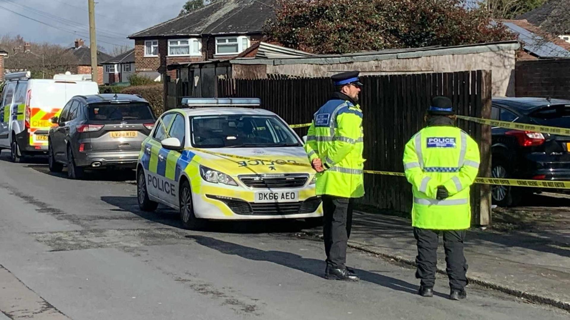 Two community support officers in yellow high-vis coats stand near a residential garage.