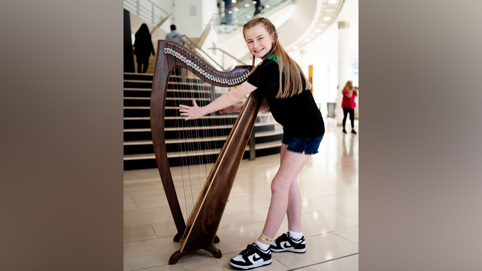 A girl is playing the harp and smiling. She's wearing a bland and green top with short and trainers. She's indoors in a large foyer with stairs behind her. People are in the background walking. 