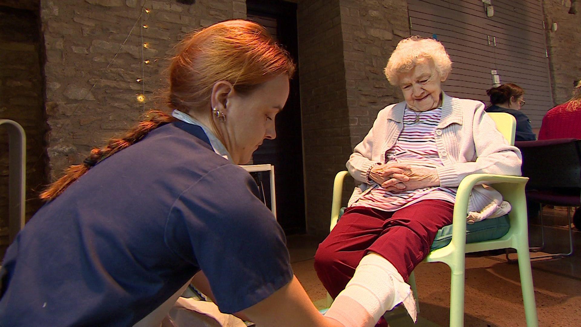 Image of a smiling elderly woman with white hair, wearing a striped t-shirt, red trousers and beige cardigan and a teddy bear pendant. She is sitting down and having her left leg dressed by a nurse who is kneeling down in front of her. The nurse is wearing a navy uniform and has red hair, tied back in a long plait. In the background can be seen two people sitting at a table.
