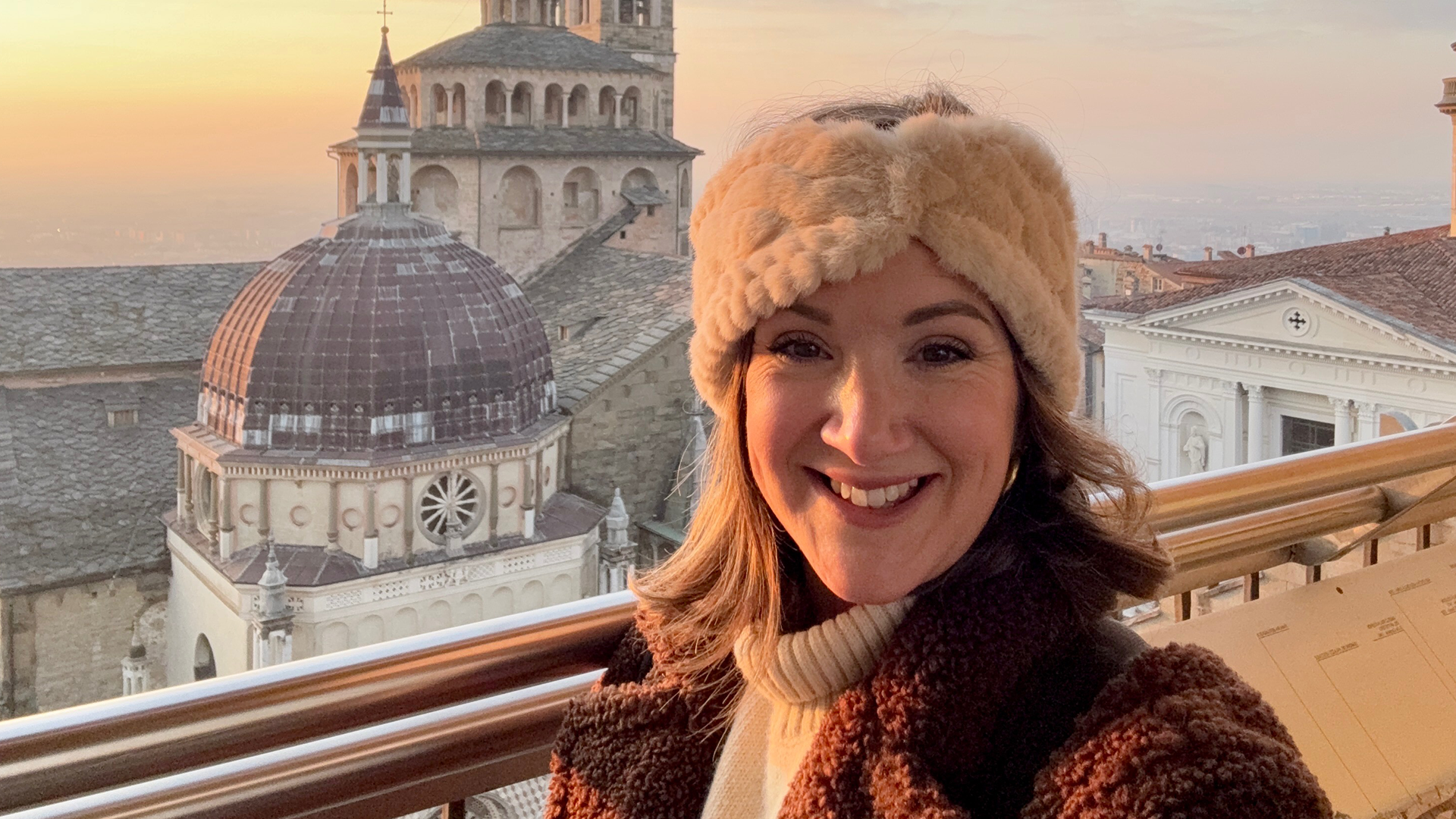 Monica Stott in front of a cathedral in Bergamo, Italy. She is smiling at the camera and wearing a cream hat and brown coat. 