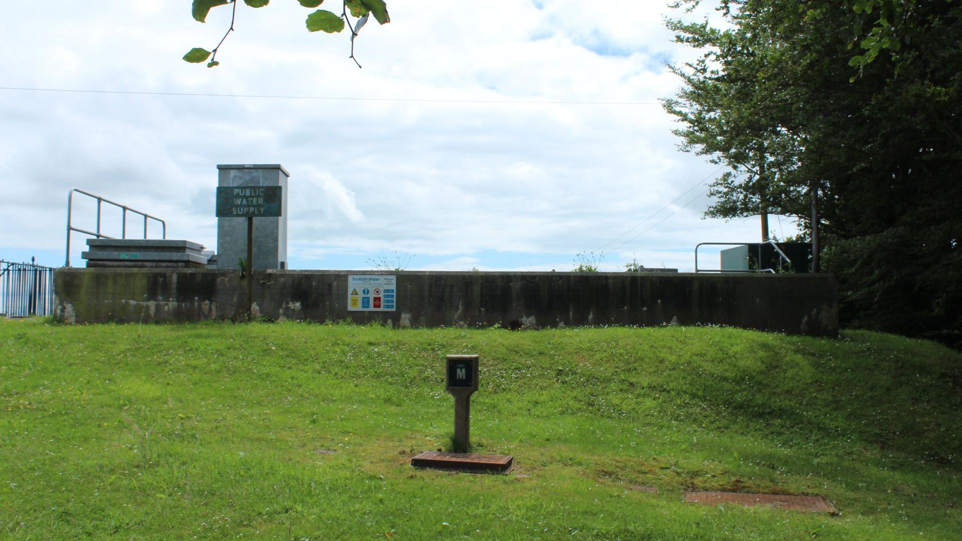 A water reservoir surrounded by grass with a "public water supply" sign in front of it